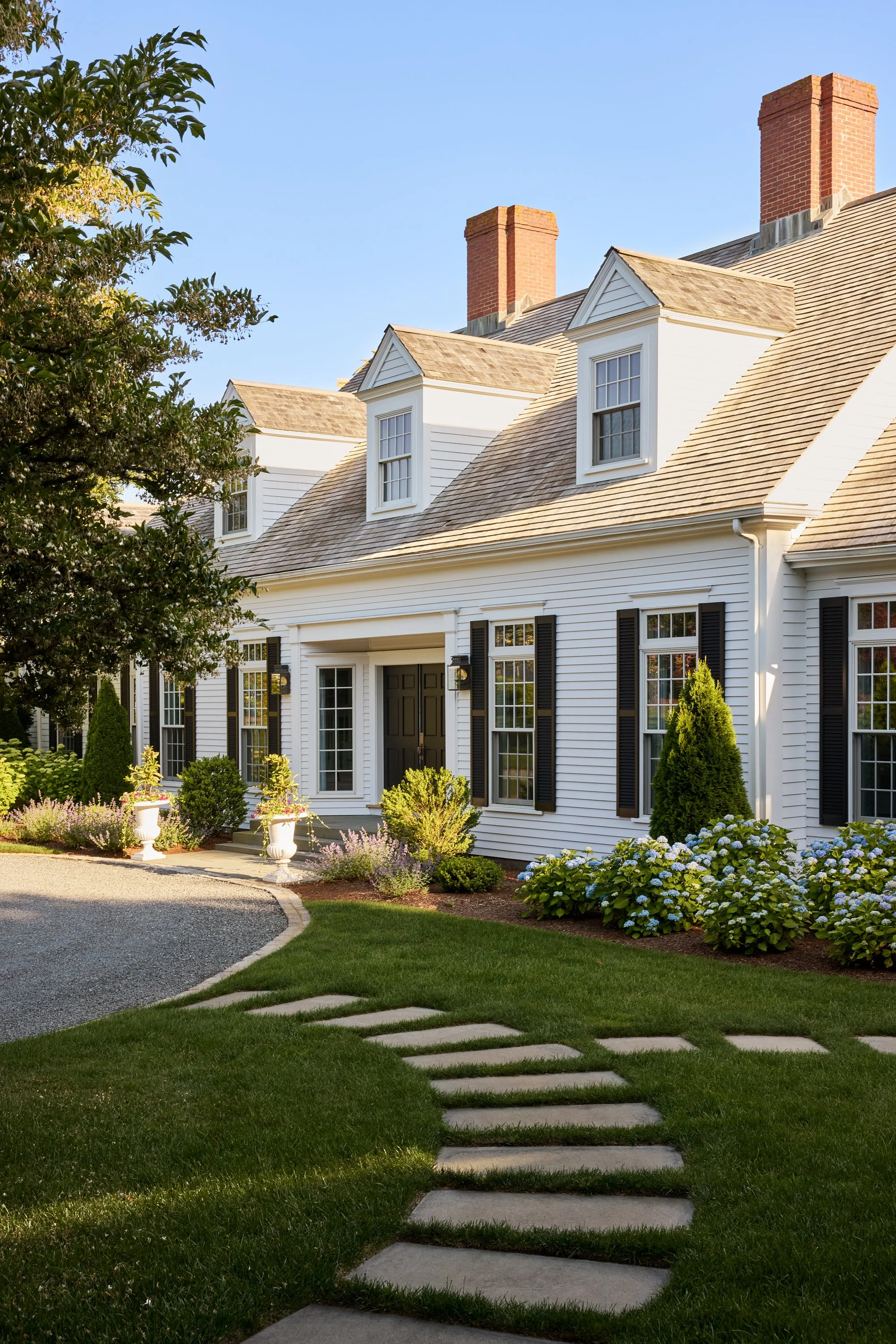Exterior view of a white house with black shutters, multiple chimneys, and dormer windows, surrounded by a landscaped garden and a stone pathway.