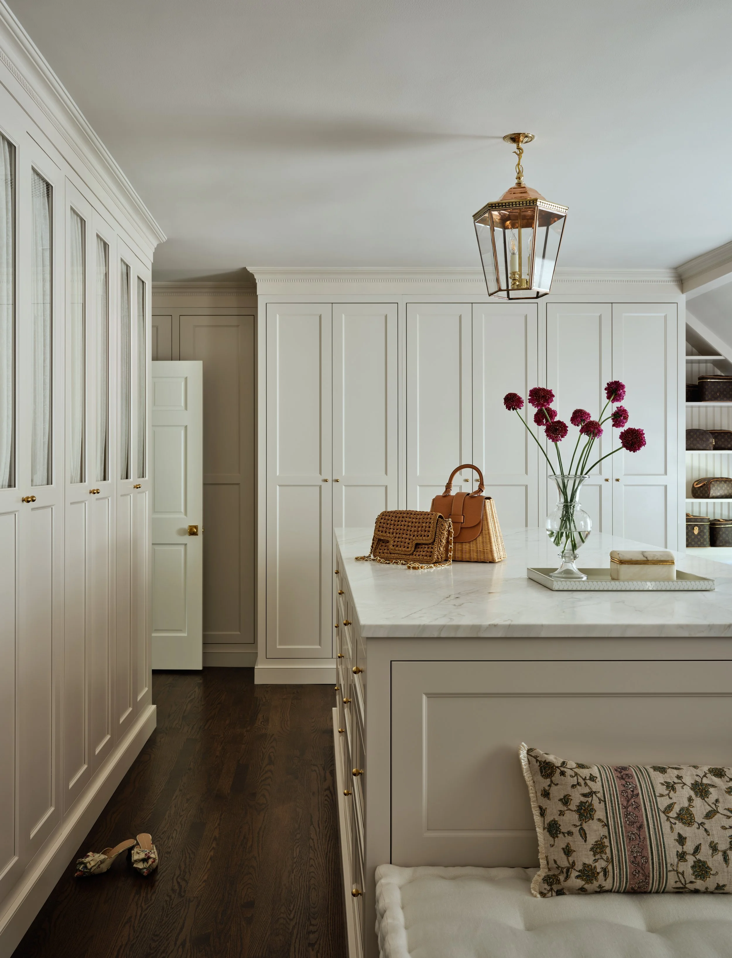 A white kitchen with a marble island, decorated with a vase of dark red flowers, handbags, and a tray, with cabinets and a hanging lantern light fixture.