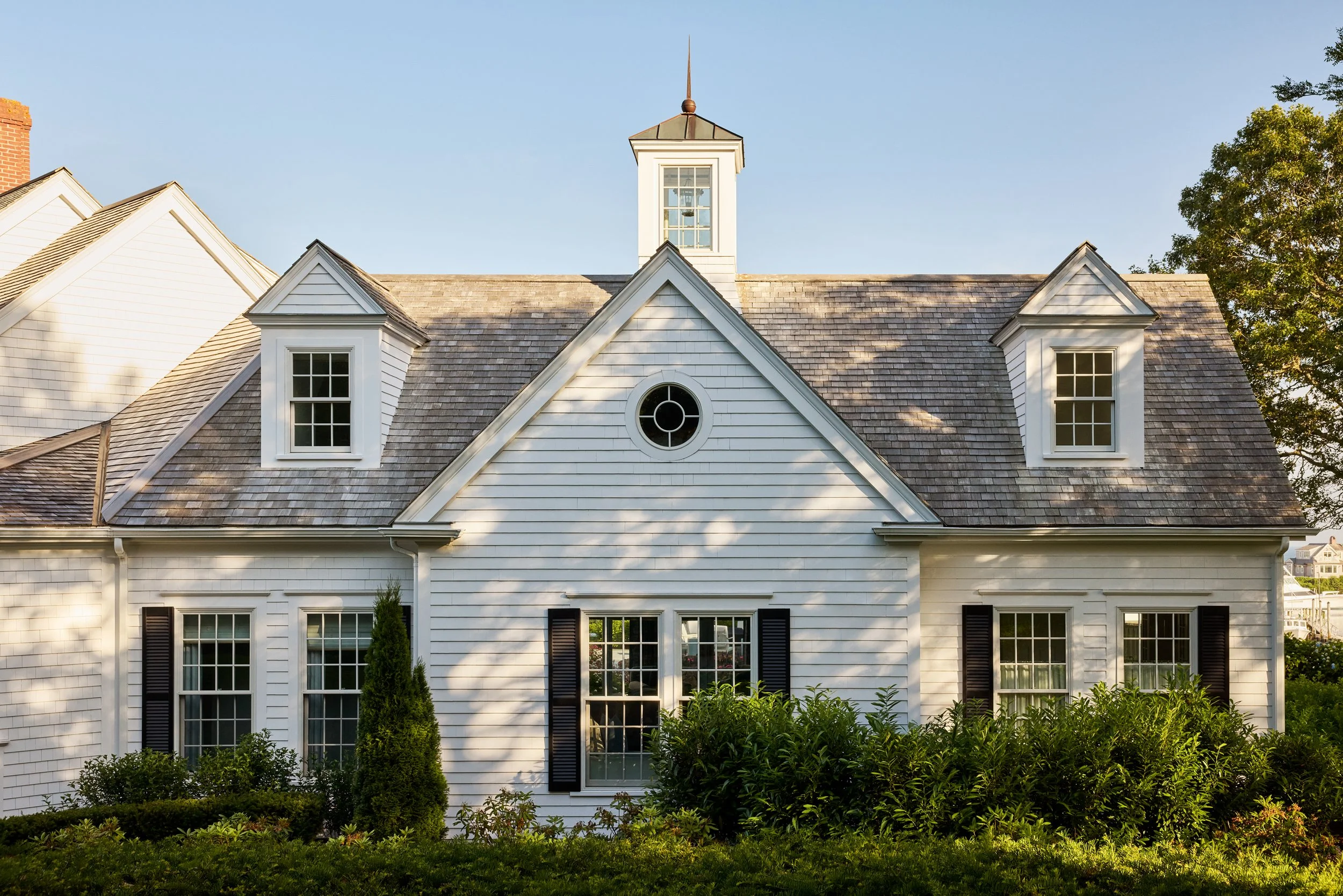 A white house with a gabled roof, black shutters, and a small bell tower on top. The house has multiple windows, some with shutters, surrounded by greenery and bushes. The sky is clear and sunny.