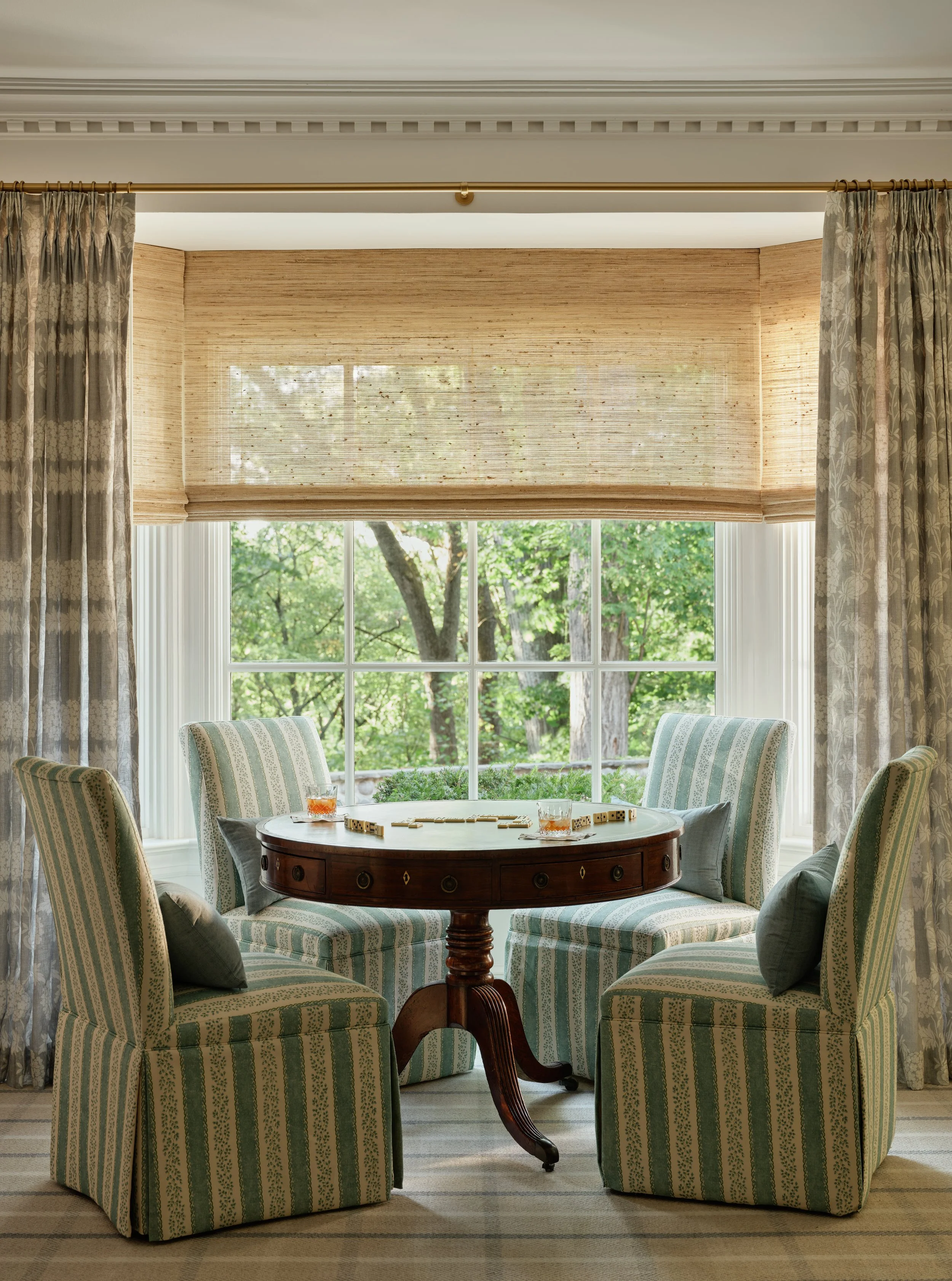 A cozy dining area with four striped upholstered armchairs around a round wooden table playing a game, set near a large window with curtains, overlooking a green outdoor scene.
