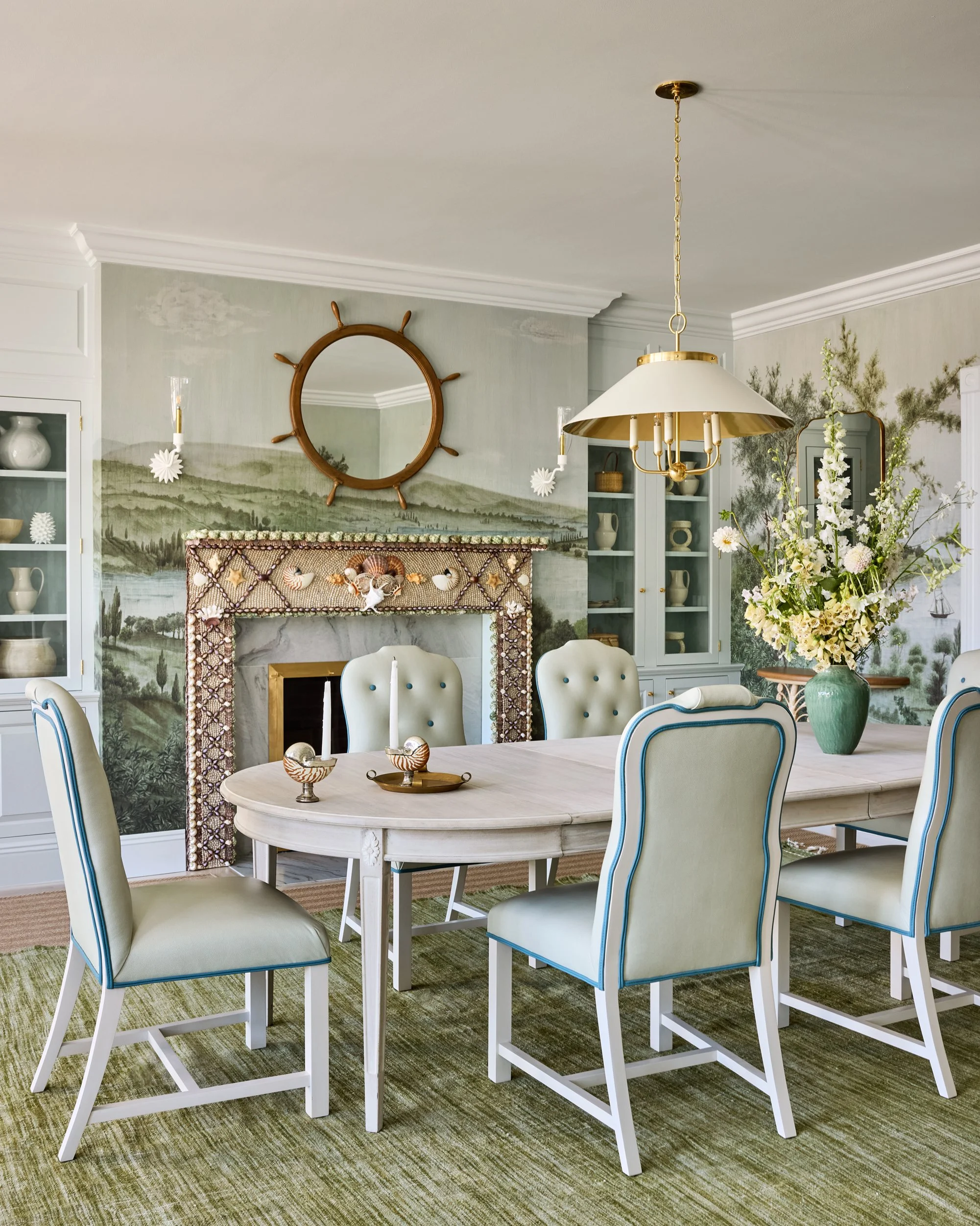 A dining room with a white oval table surrounded by six cream-colored chairs with blue piping, a large vase of white flowers, a mural of a landscape on the wall, a fireplace decorated with seashells, a gold chandelier, and built-in white cabinets wit