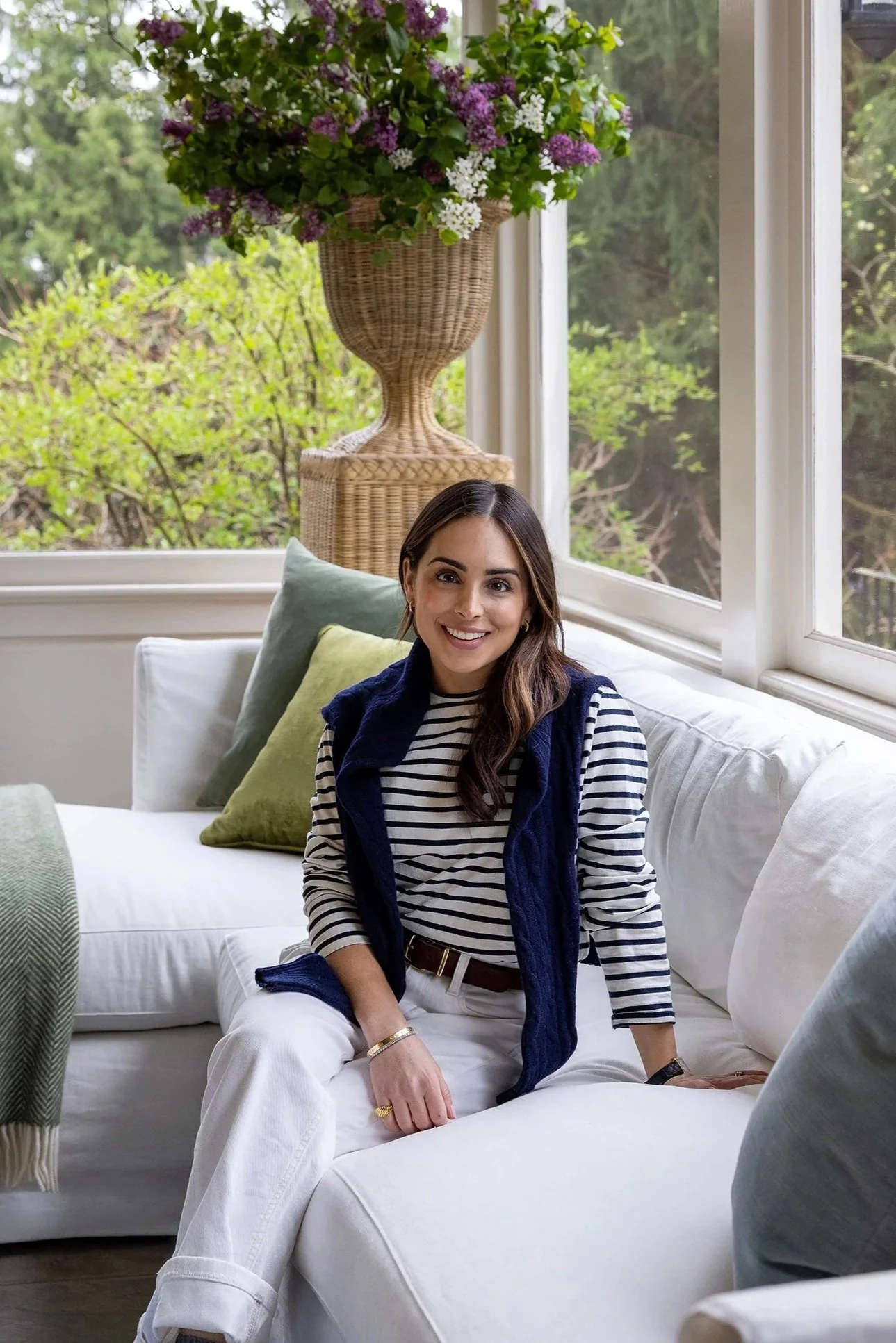 Smiling woman sitting on a white sofa in a bright room with large windows, green pillows, and a tall wicker vase with purple and white flowers behind her.