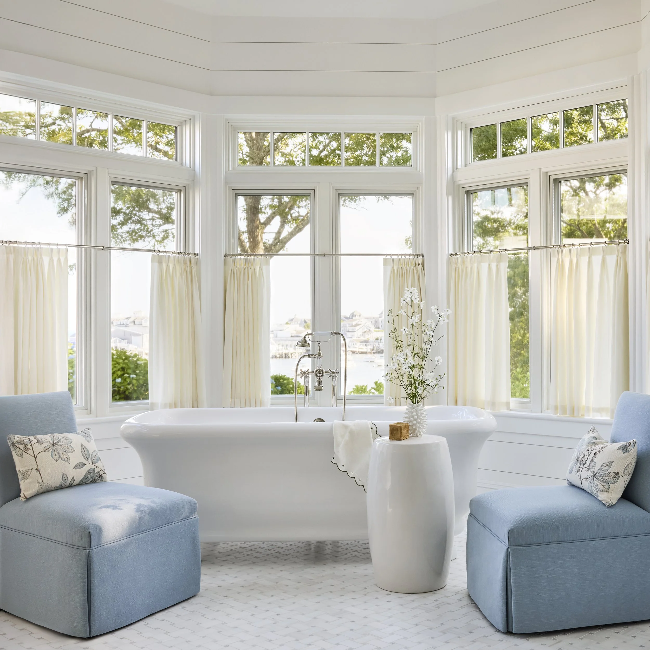 Bright bathroom with a white clawfoot bathtub, two light blue armchairs with patterned pillows, a white vase with flowers, and large windows with cream curtains showing trees and water outside.