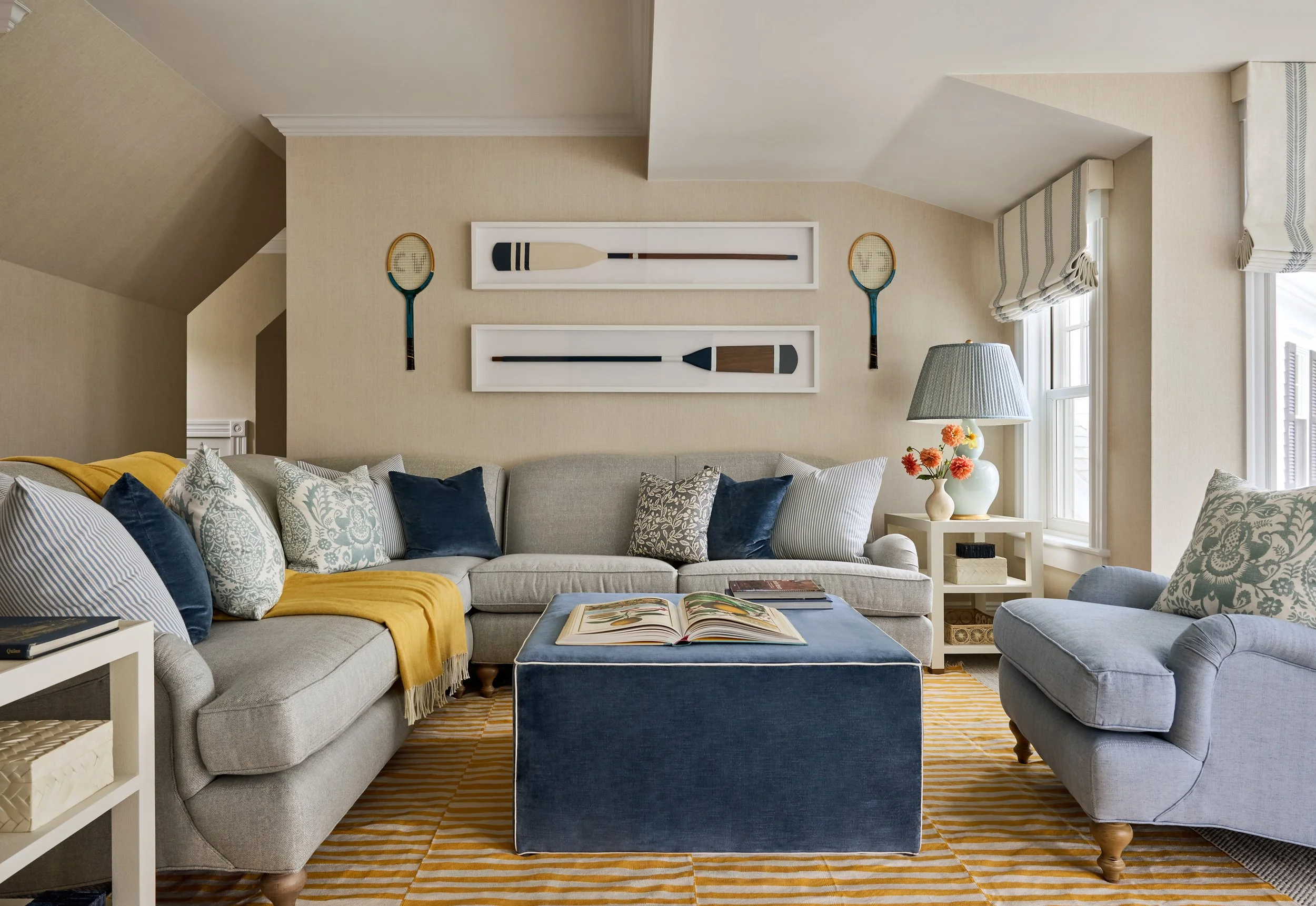 Cozy living room with a light gray sofa, blue ottoman, light gray armchair, yellow throw blanket, assorted decorative pillows, tropical art book on the ottoman, striped rug, side table with books, ceramic vase with flowers, wicker lamp, and tennis ra