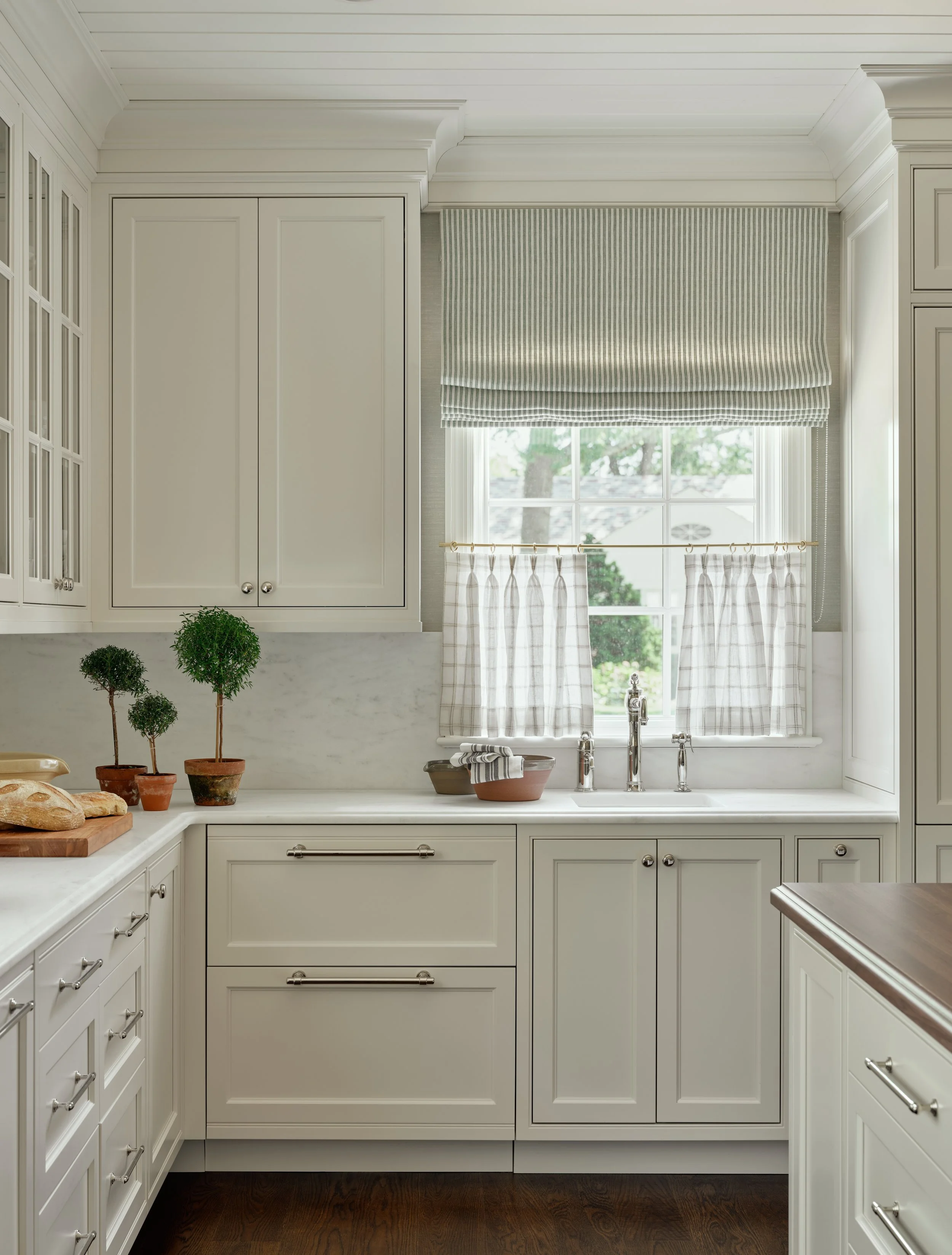 A bright kitchen with white cabinets, a window with checkered curtains, and a marble countertop with small potted trees and bread.
