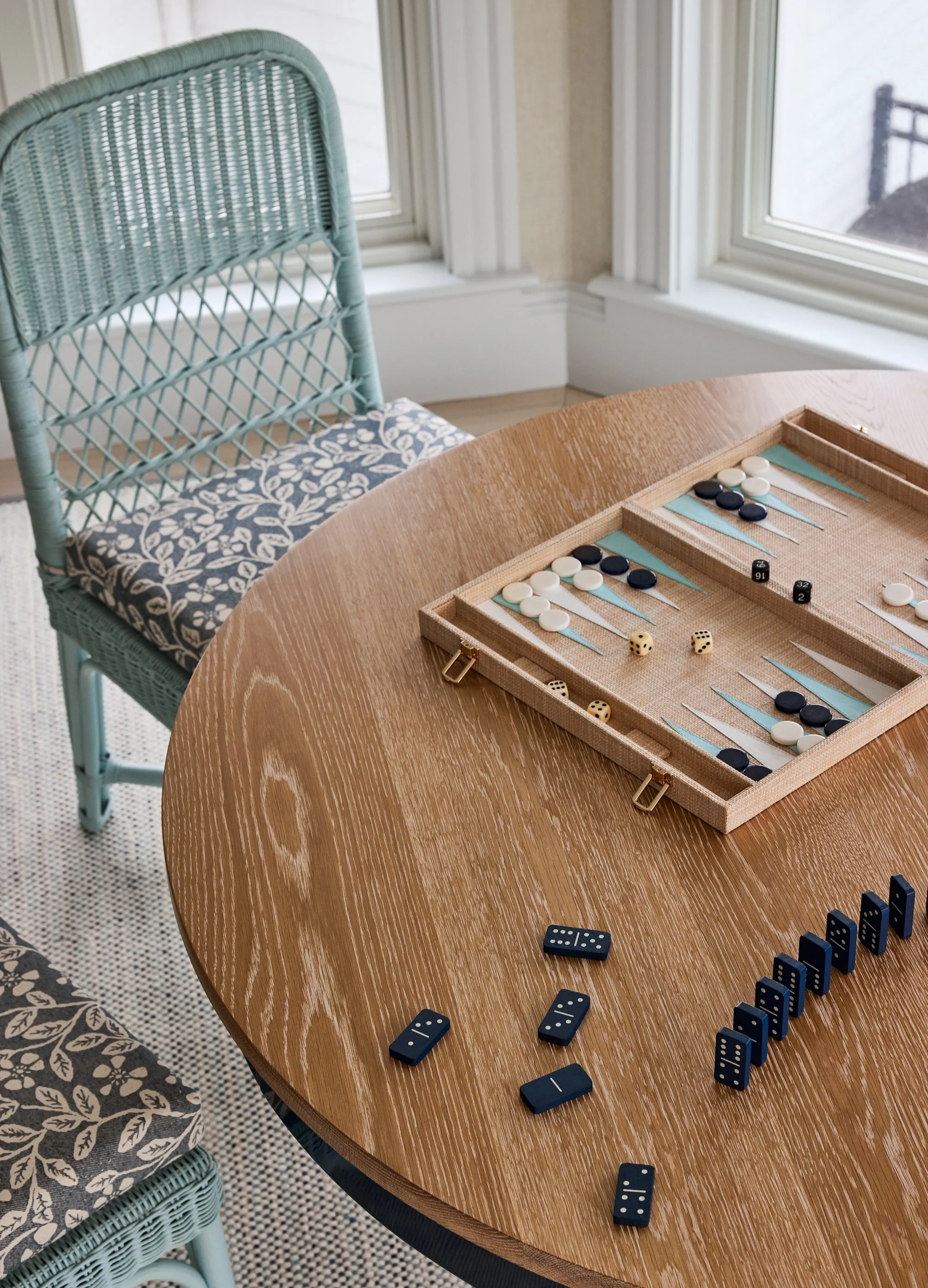 A wooden table with a backgammon set, dominoes, and a line of domino pieces, surrounded by chairs, next to windows in a sunlit room.