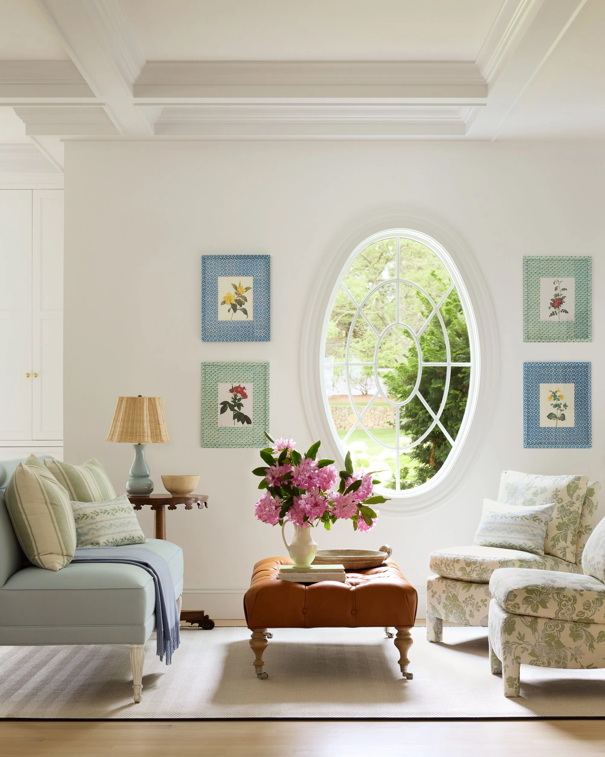 A cozy living room with a large oval window, a sofa with cushions, a floral armchair, a side table with a lamp, a vase of pink flowers on an ottoman, and framed botanical art on the wall.