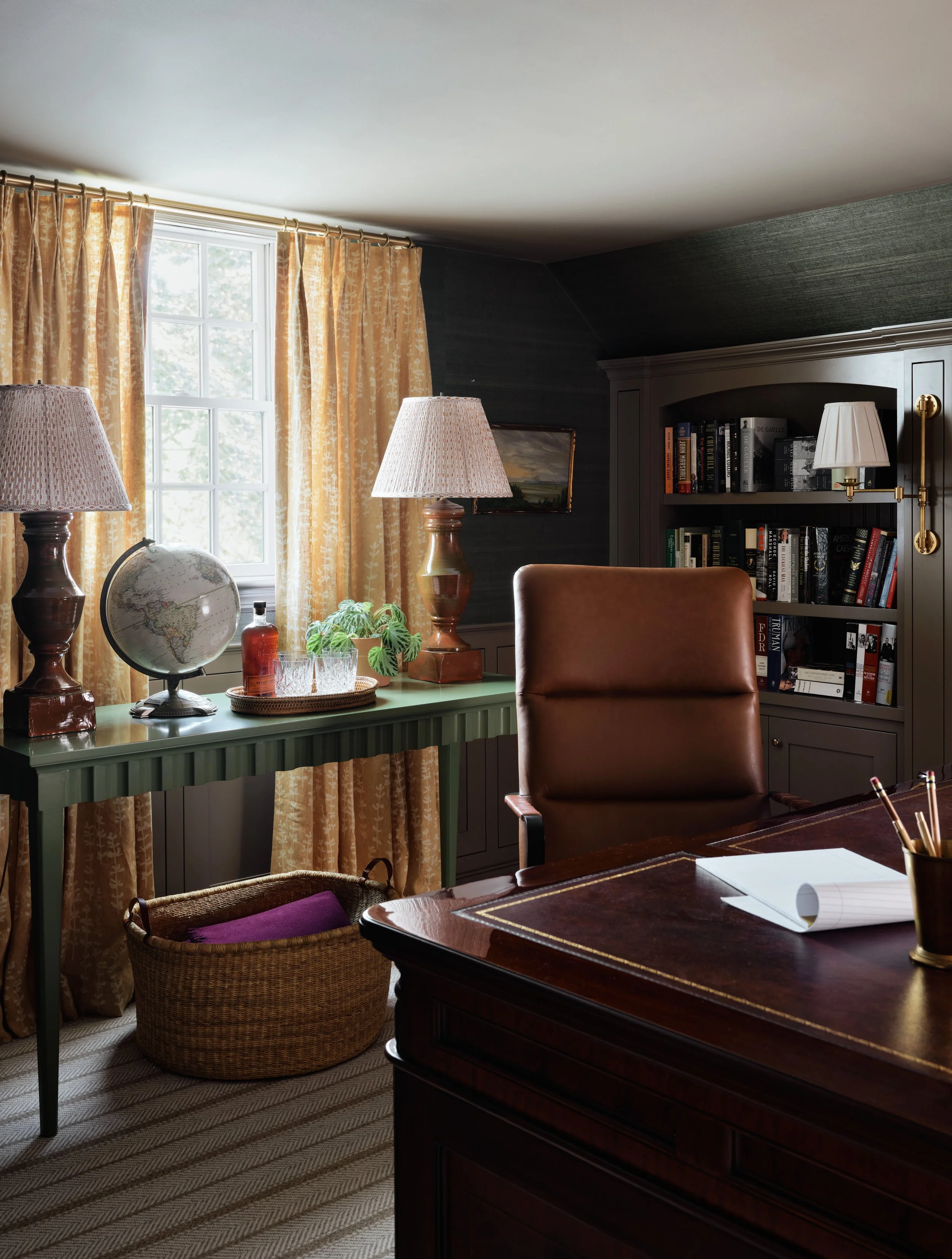 A cozy, well-lit home office with a large window, yellow curtains, a green desk, a globe, a plant, a brown leather chair, a dark wooden bookshelf with books, a table lamp, and a dark wood desk with writing utensils.