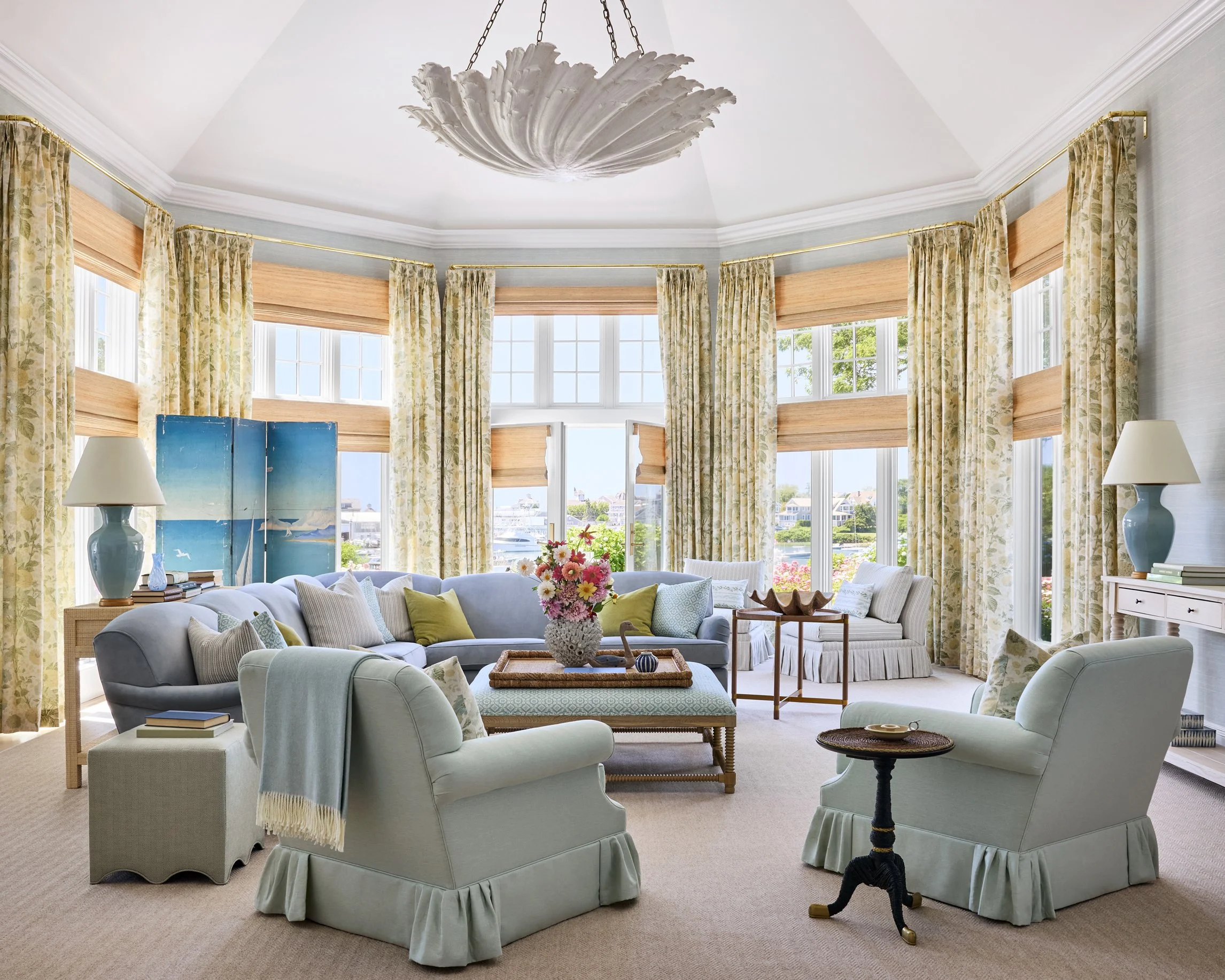Bright living room with large bay windows, pastel-colored sofas, armchairs, floral curtains, a floral centerpiece, and decorative lamps.