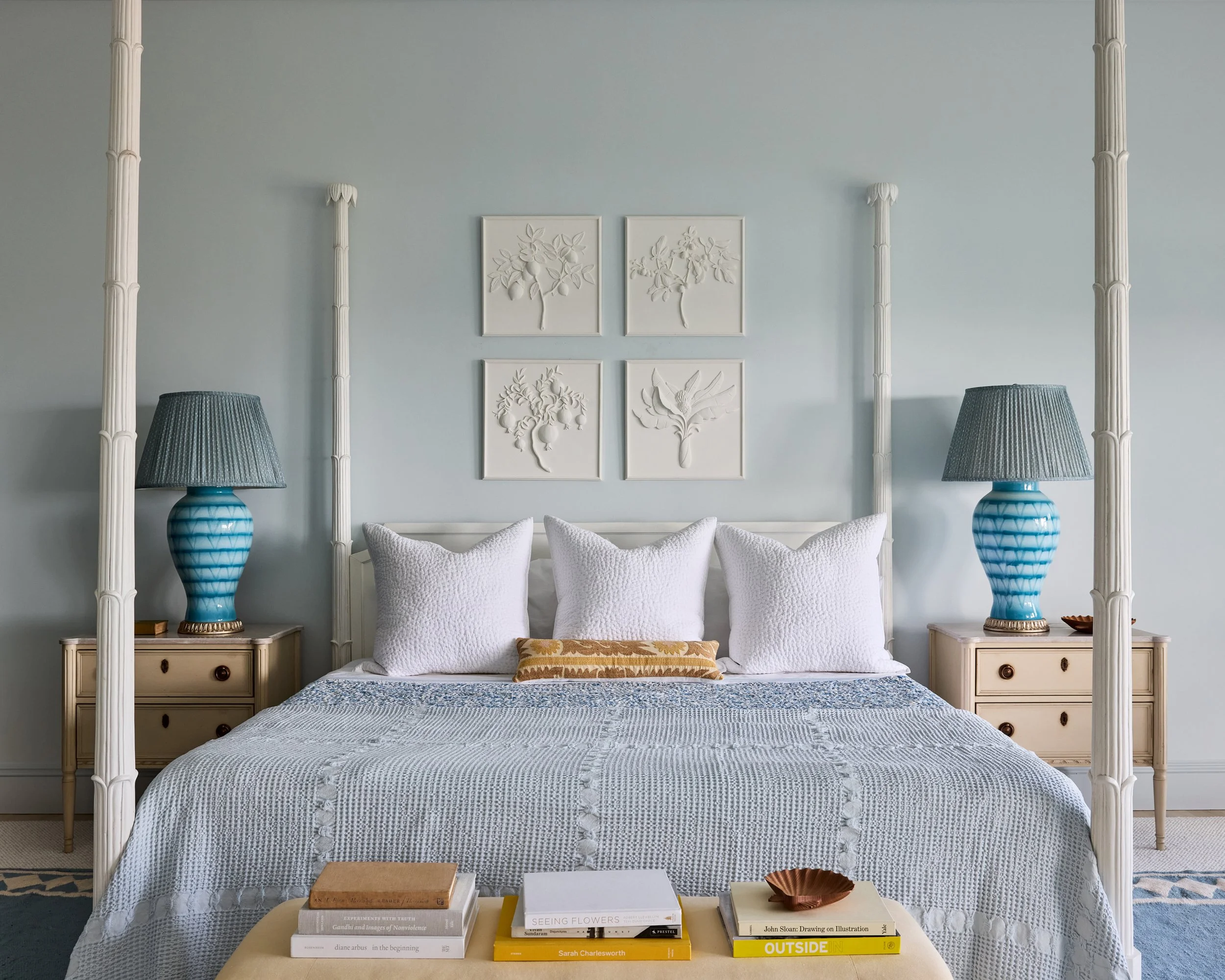 A cozy bedroom with a four-poster bed, white bedding, four large white pillows, and a decorative pillow. Two matching blue lamps with dark shades are on cream nightstands on each side of the bed. Above the bed, four white art pieces with raised botan