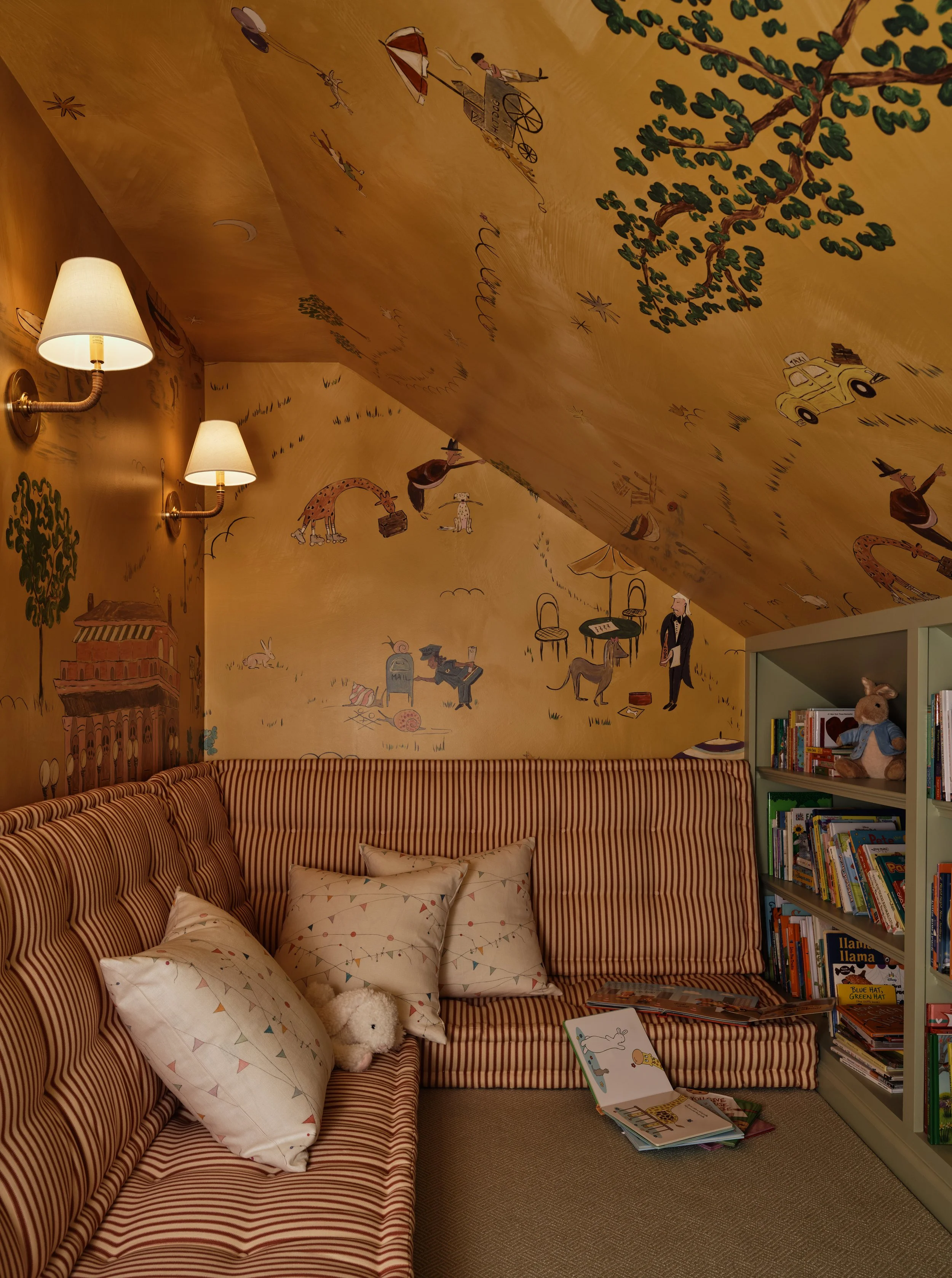 A cozy room with striped red and white couch, pillows, bookshelves filled with children's books, and whimsical wall and ceiling illustrations of animals, trees, and people.