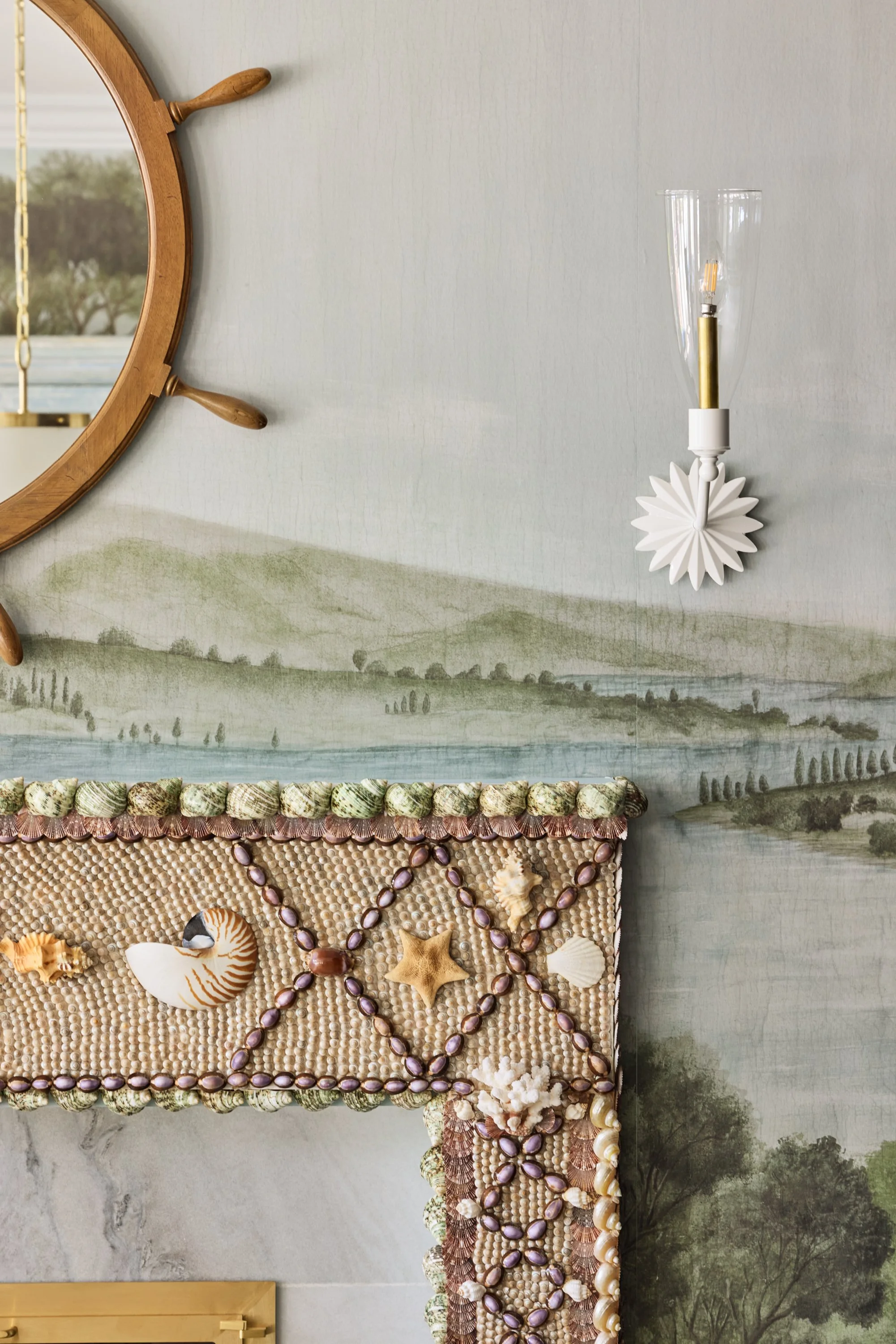 Decorative wall with ship wheel mirror, shell art, chandelier, and mural of landscape.