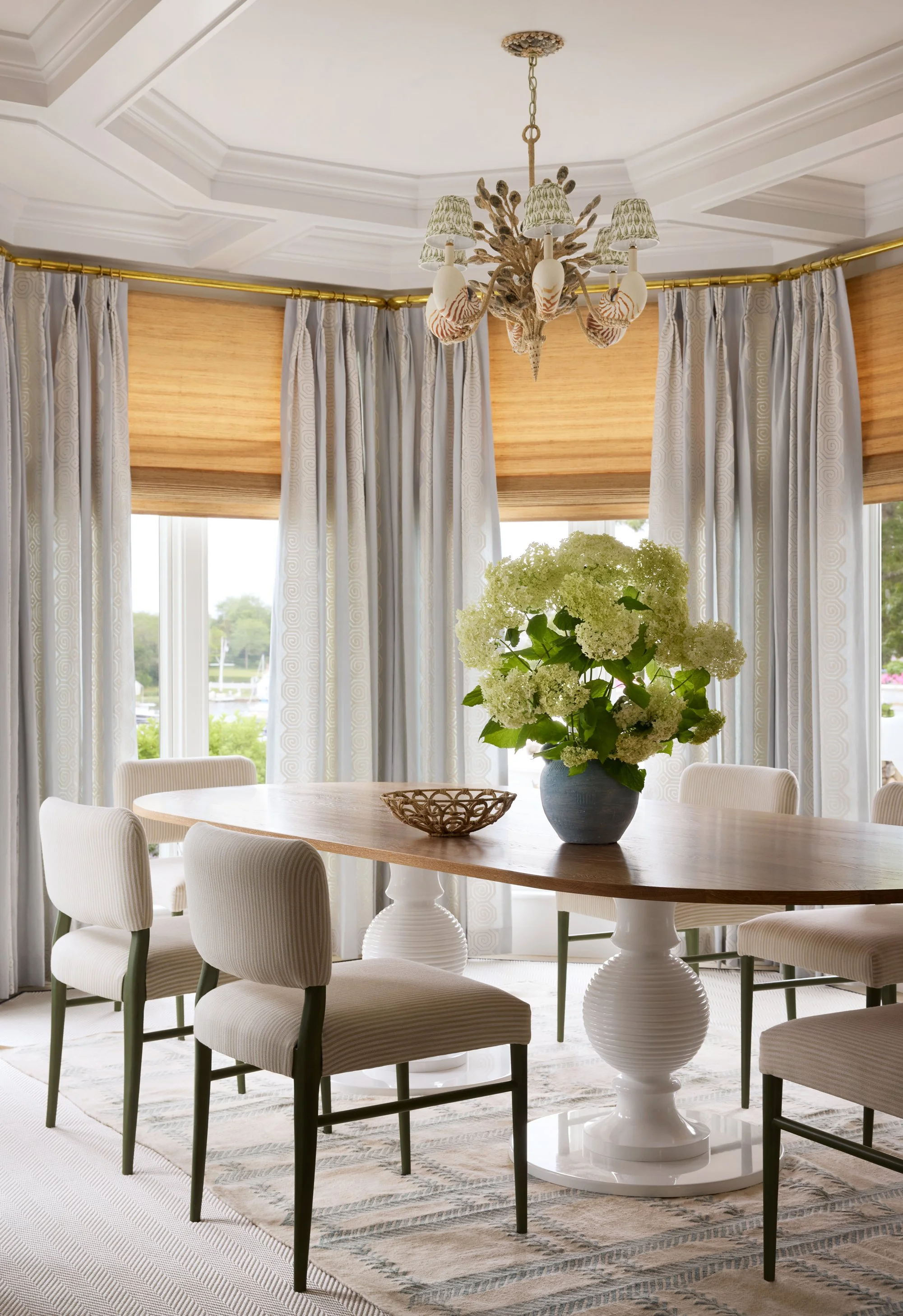 A dining room with a wooden oval table, six upholstered chairs, a large flower arrangement on the table, a decorative chandelier hanging from the ceiling, and windows with white curtains and wooden blinds.