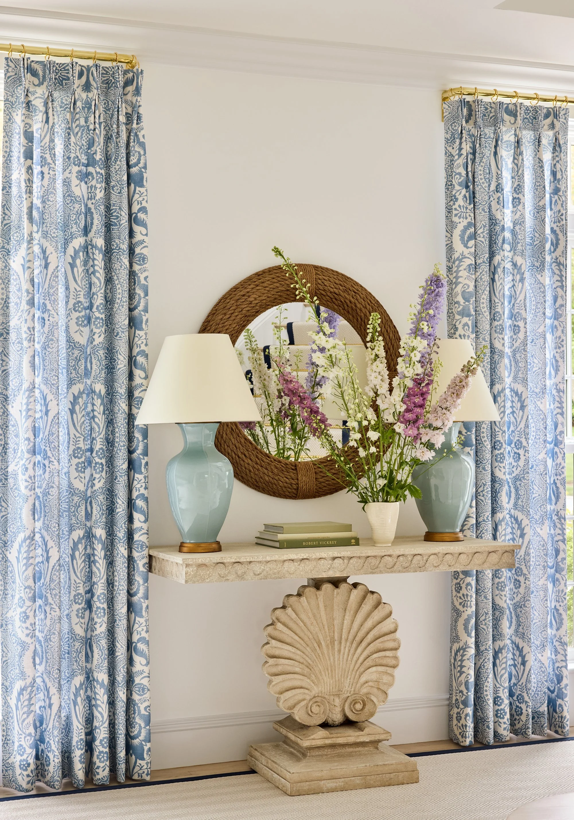 A decorative interior entryway with blue and white patterned curtains, a stone console table with a shell-shaped base, two pale blue lamps, a white vase with purple and white flowers, and a large circular mirror with a woven frame.