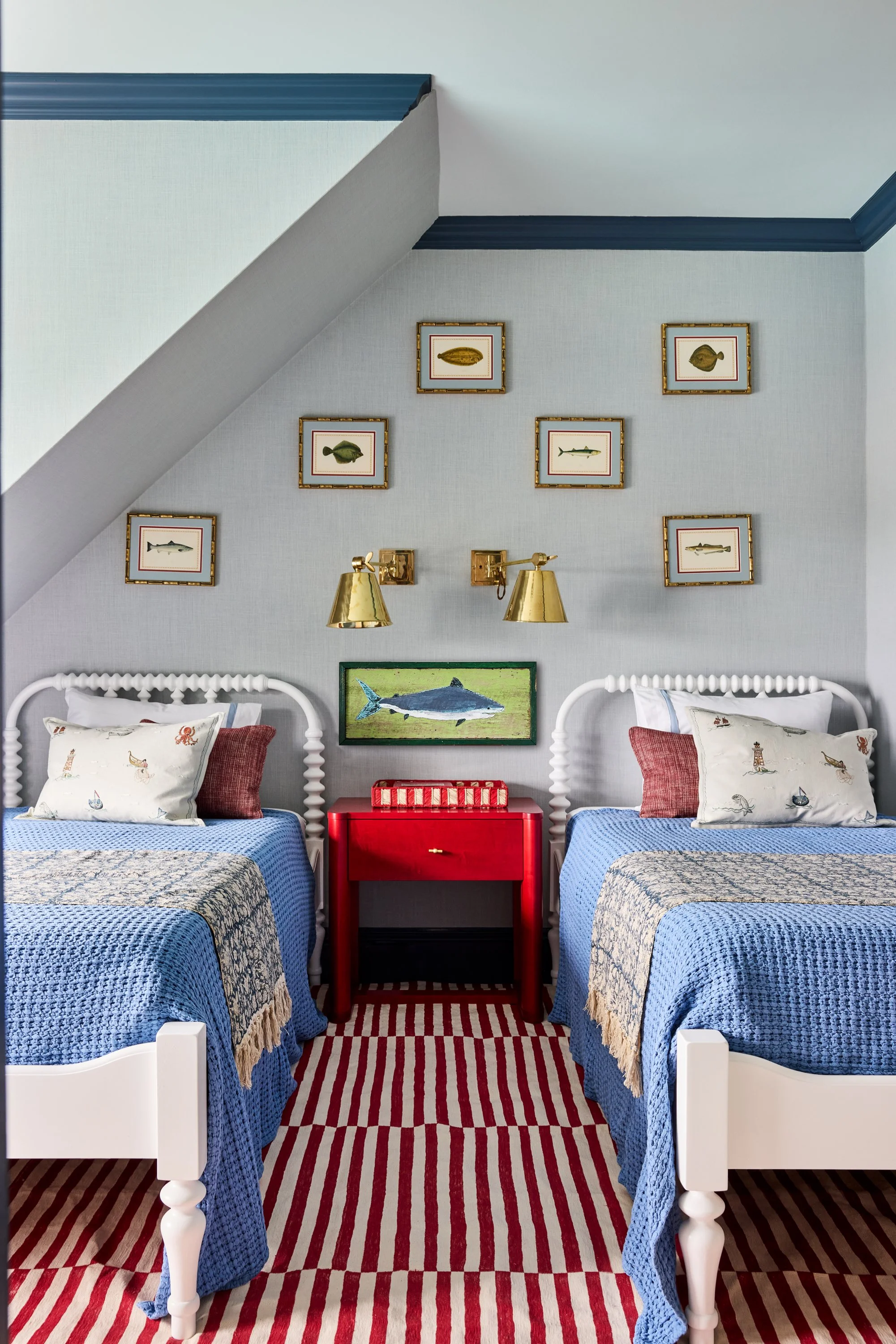 A bedroom with two white beds featuring nautical-themed bedding, a red nightstand between the beds, and a wall decorated with framed fish prints and a painting of a shark, set under a sloped ceiling with blue trim.