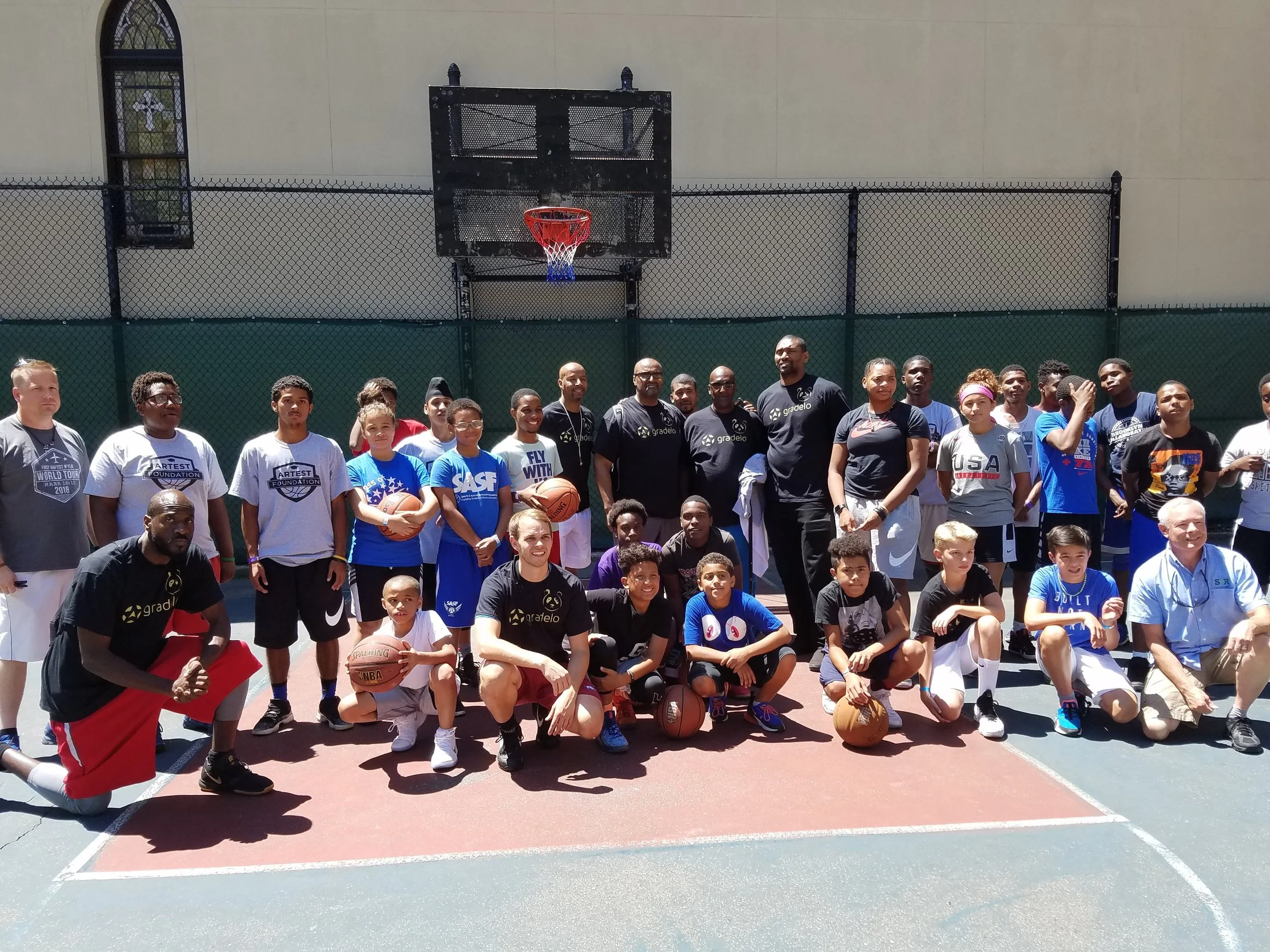 Group of people, including children and adults, on a basketball court, some holding basketballs, with a basketball hoop and backboard in the background.