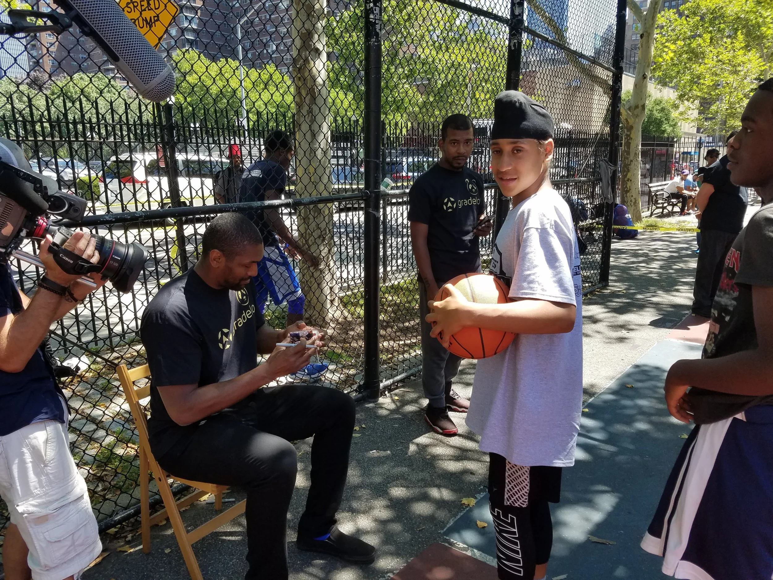 A group of young men, some in sportswear, gather on an outdoor basketball court near a chain-link fence. One young man in a gray shirt and black cap holds a basketball, smiling at the camera. Another is seated, signing an autograph, with a camera per