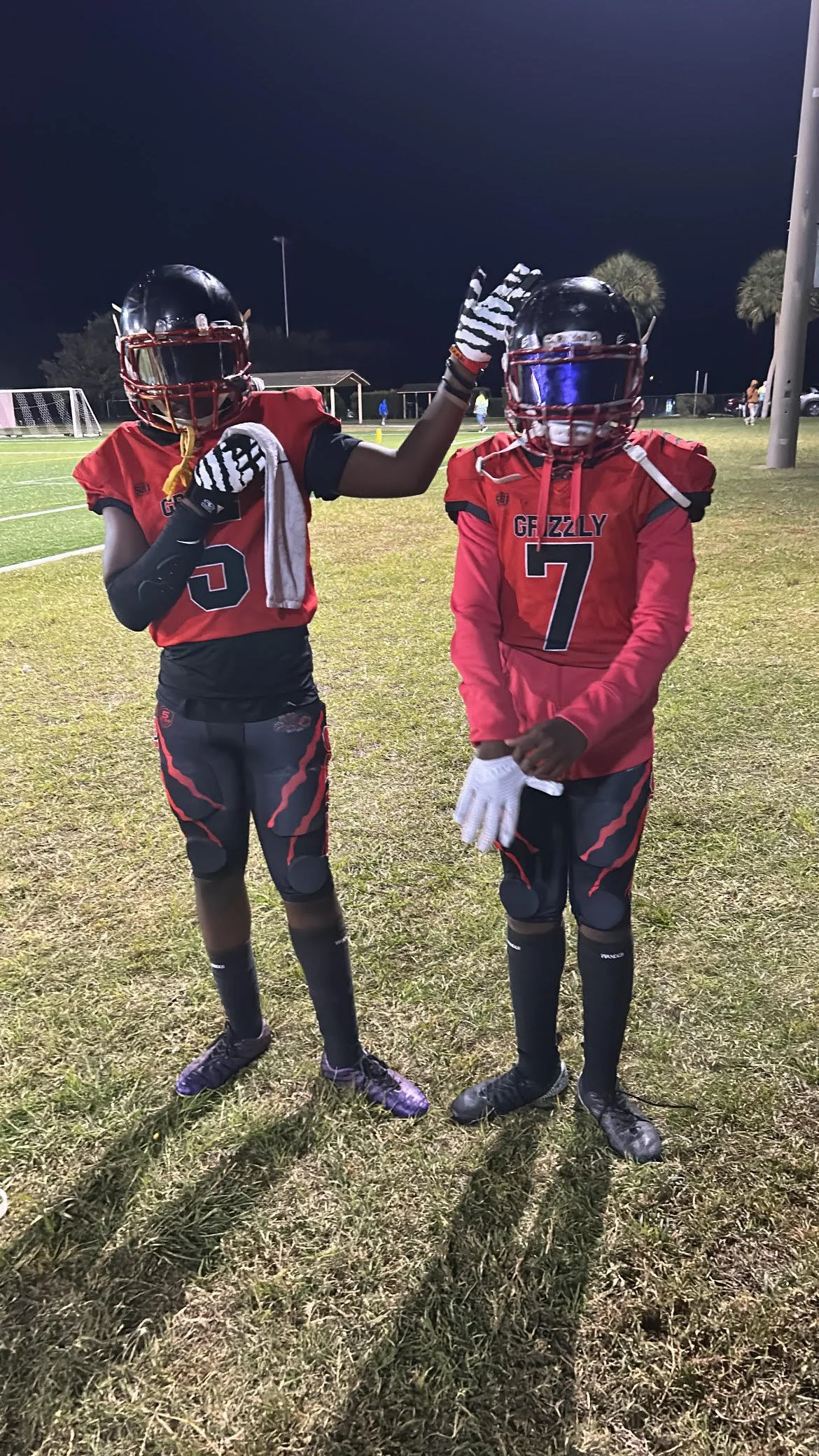 Two young football players in red and black uniforms, wearing helmets with face shields, standing on a football field at night. One player is giving a high-five to the other.
