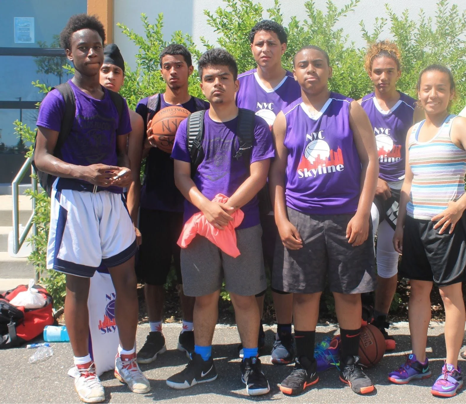 Group of young basketball players and a coach outside, some wearing purple jerseys with 'NYC Skyline' logo, holding basketballs with sports gear on ground and backpacks nearby.