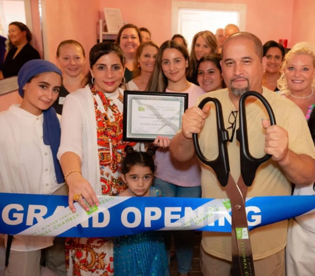 Group of people celebrating grand opening, holding a certificate, with a man holding large scissors, woman with a child, and a ribbon across the foreground.