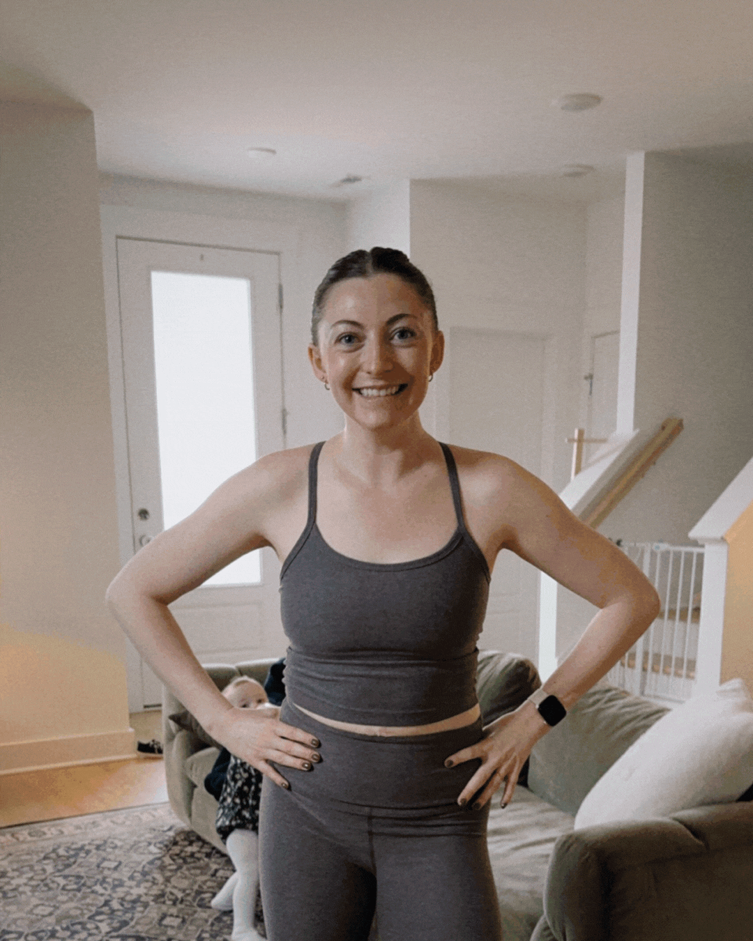 Woman in workout clothes smiling with hands on her hips in a living room.