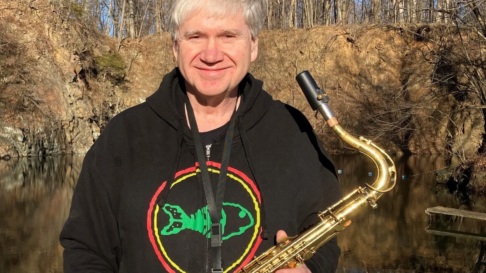 A smiling man with gray hair holding a saxophone outdoors near a body of water, with bare trees in the background during a sunny day.