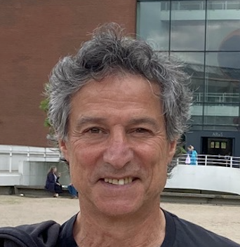 A man with curly gray hair smiling outdoors with a modern building in the background.