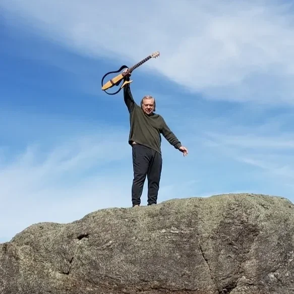 Man standing on a large rock formation holding a guitar in the air against a cloudy sky.