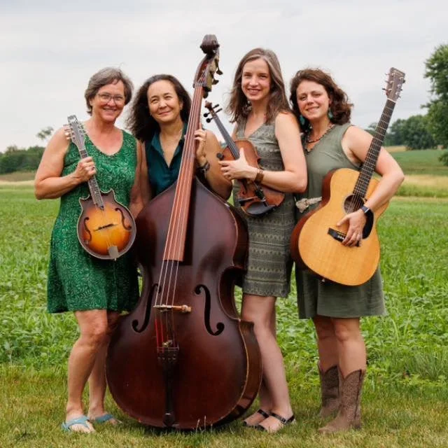 Four women standing outdoors in a grassy field, each holding a musical instrument, smiling at the camera.
