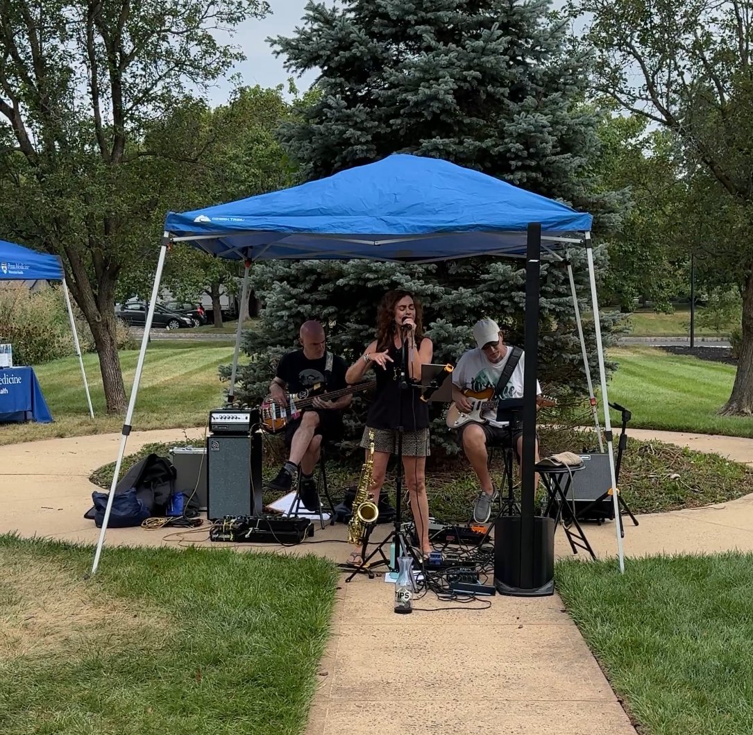 A band performing outdoors under a blue canopy tent, with a woman singing into a microphone flanked by two guitar players, on a sidewalk in a park with green grass and trees.