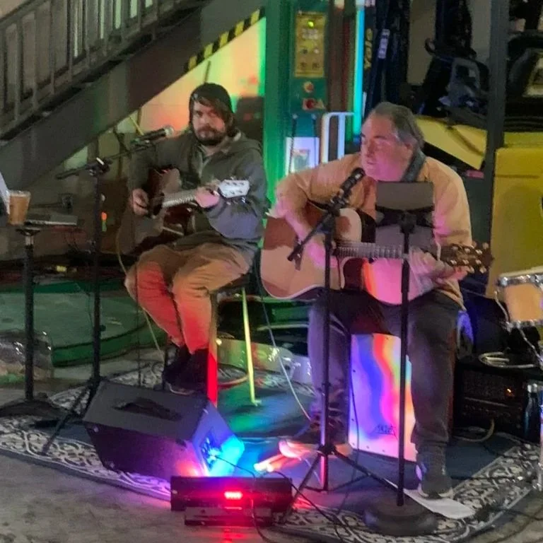 Two musicians playing acoustic guitars and singing into microphones, performing on a small stage in a dimly lit venue with colorful lighting.