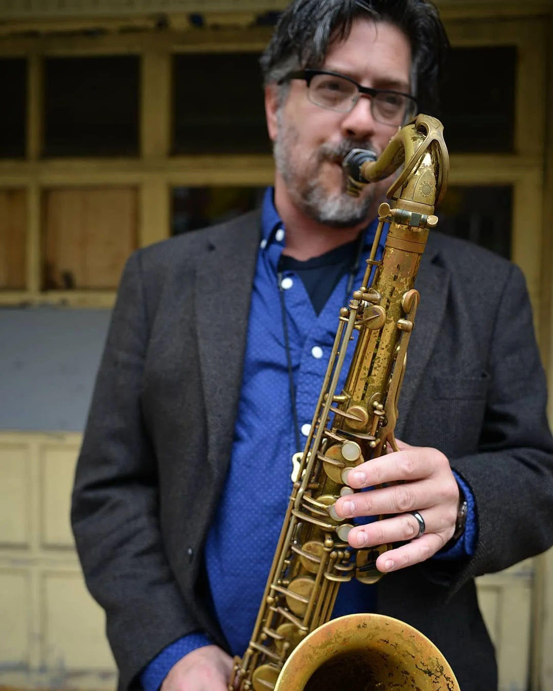 A man with glasses and a beard playing a gold-colored saxophone indoors.