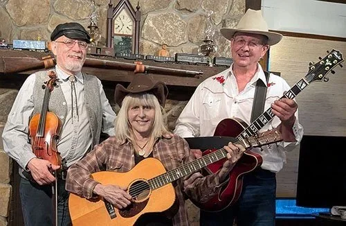 Three musicians, two men and one woman, performing together in a cozy indoor setting. The man on the left is playing a violin, the woman in the center is playing an acoustic guitar, and the man on the right is playing an electric guitar and wearing a cowboy hat. They are smiling.