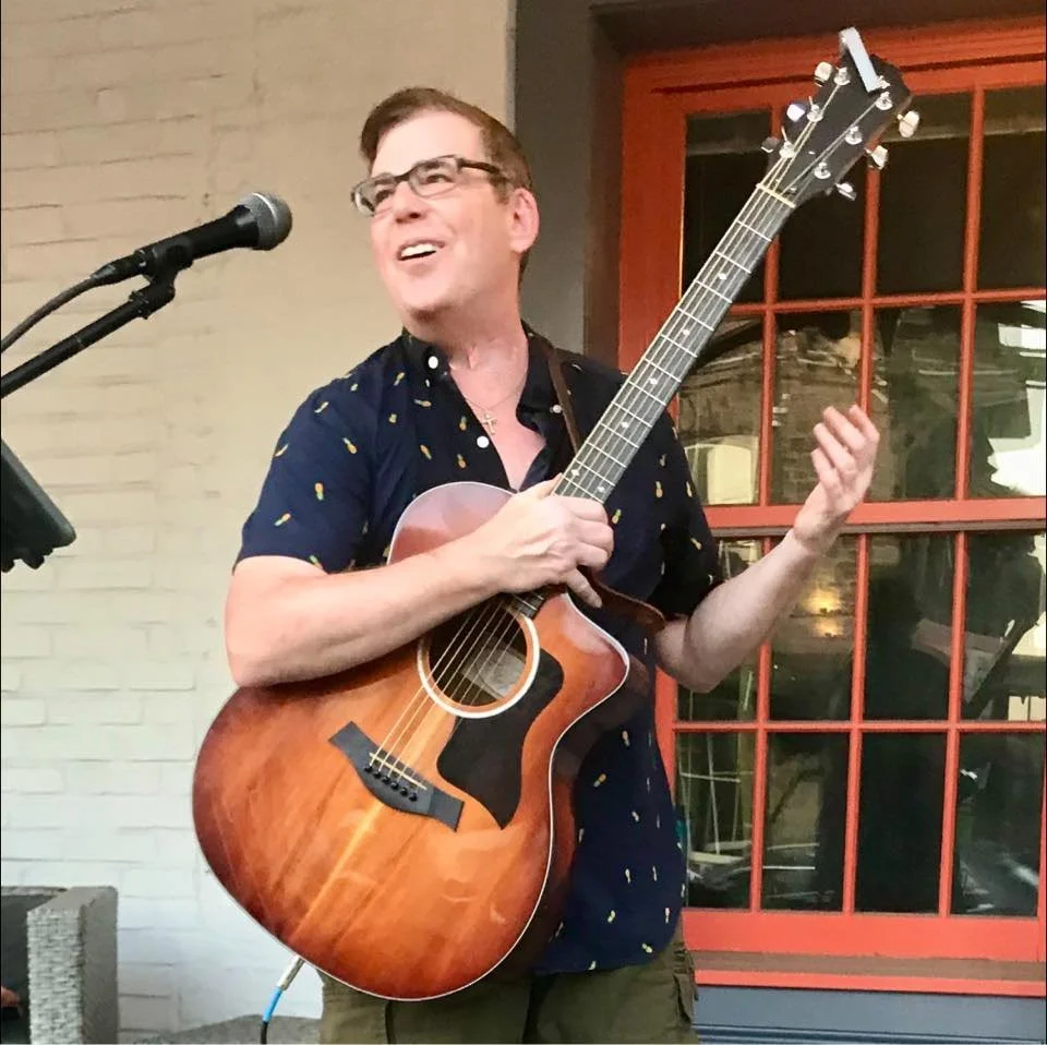A man singing into a microphone while playing an acoustic guitar indoors near a brick wall and a red window.