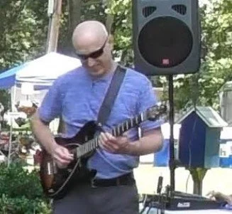 Man wearing sunglasses playing an acoustic guitar outdoors with a speaker in the background.