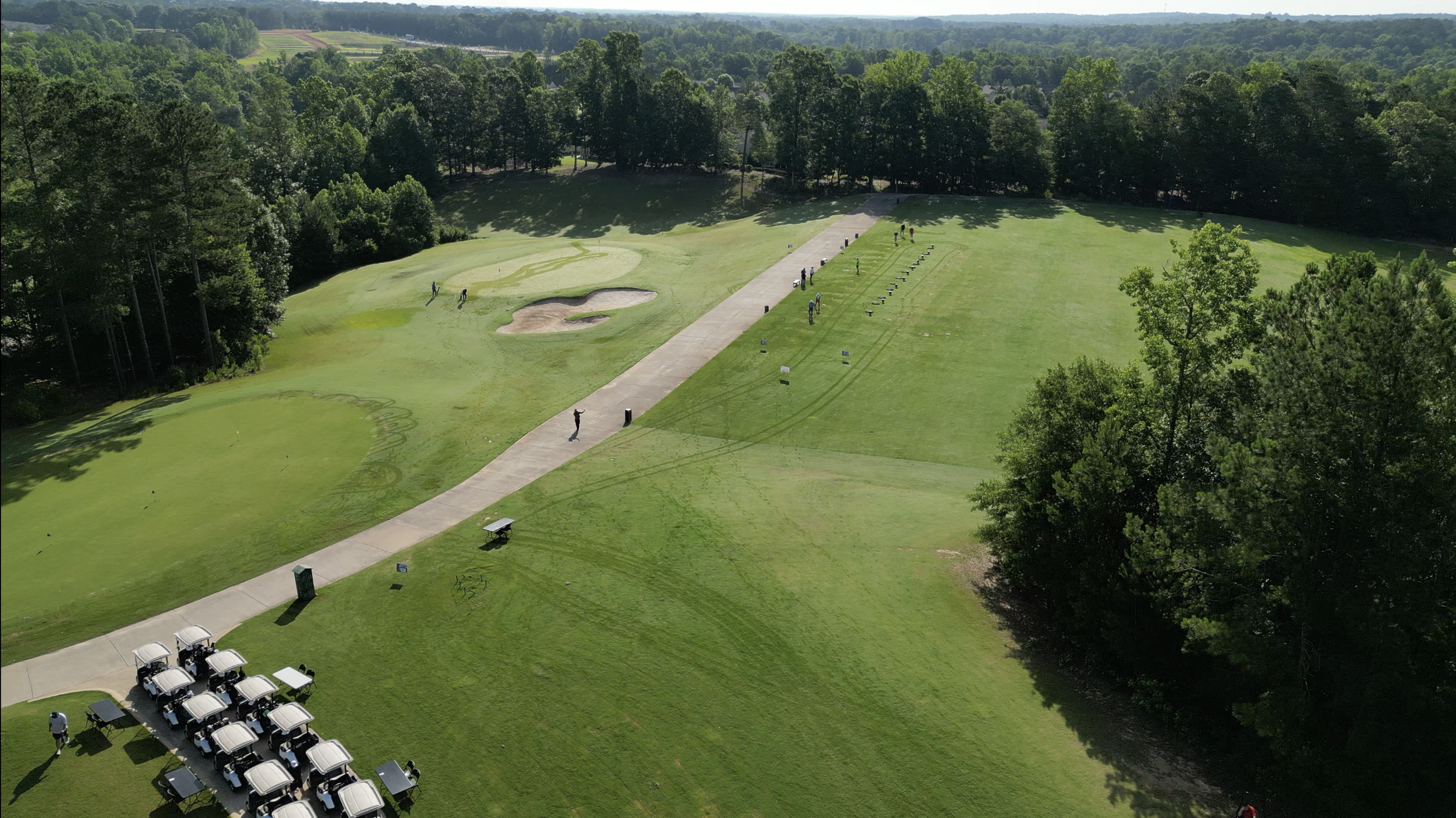 An aerial view of a golf course with golf carts parked on the left, a pathway with people walking, and a grassy area with flags and equipment, surrounded by trees.