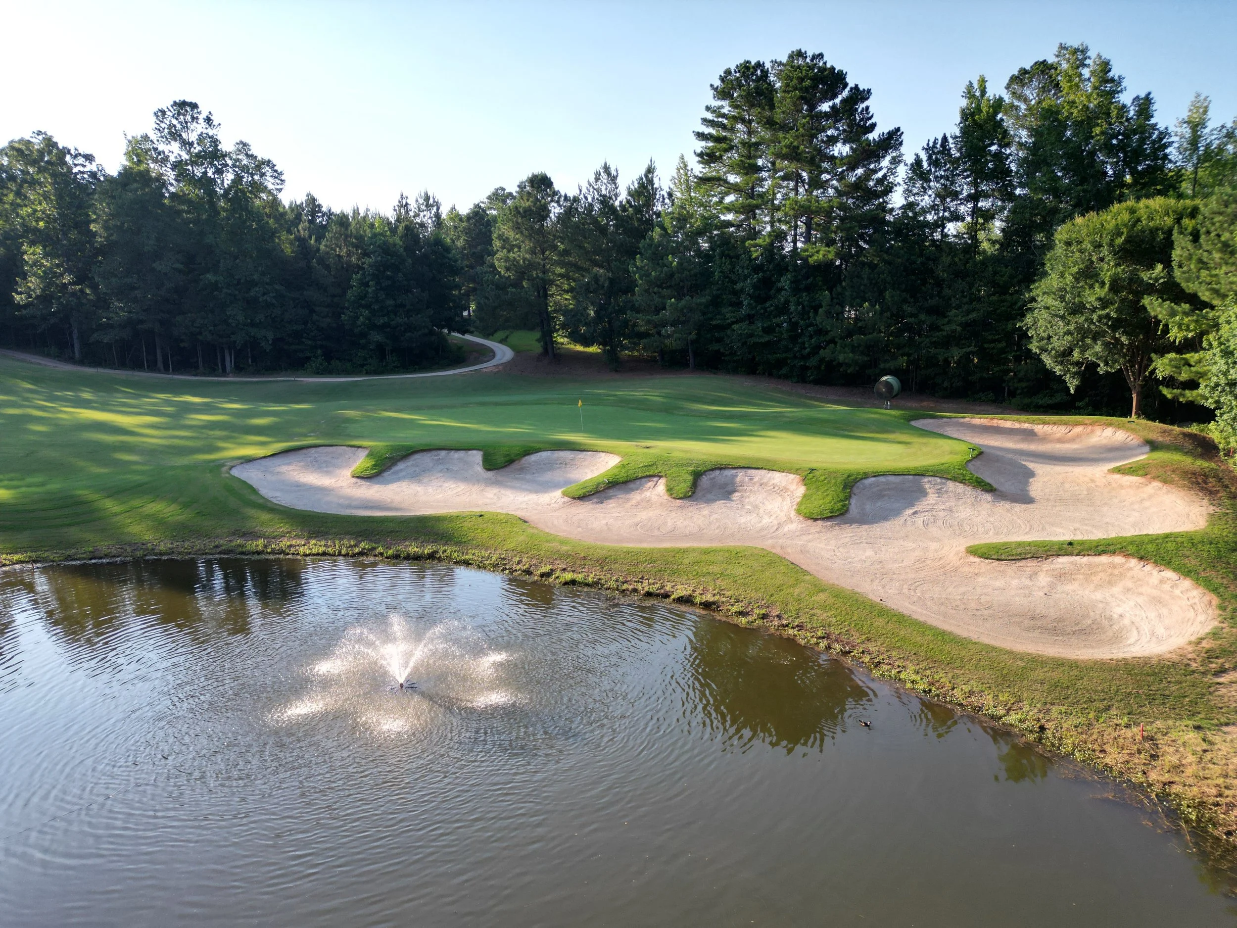 Golf course with green, sand bunkers, a flag, and a pond with a fountain, surrounded by trees.