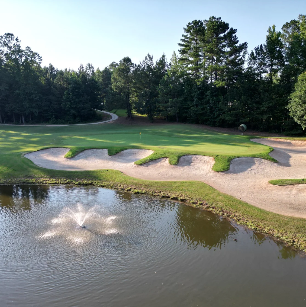 A golf course with a water hazard, sand bunkers, a putting green, and trees in the background.