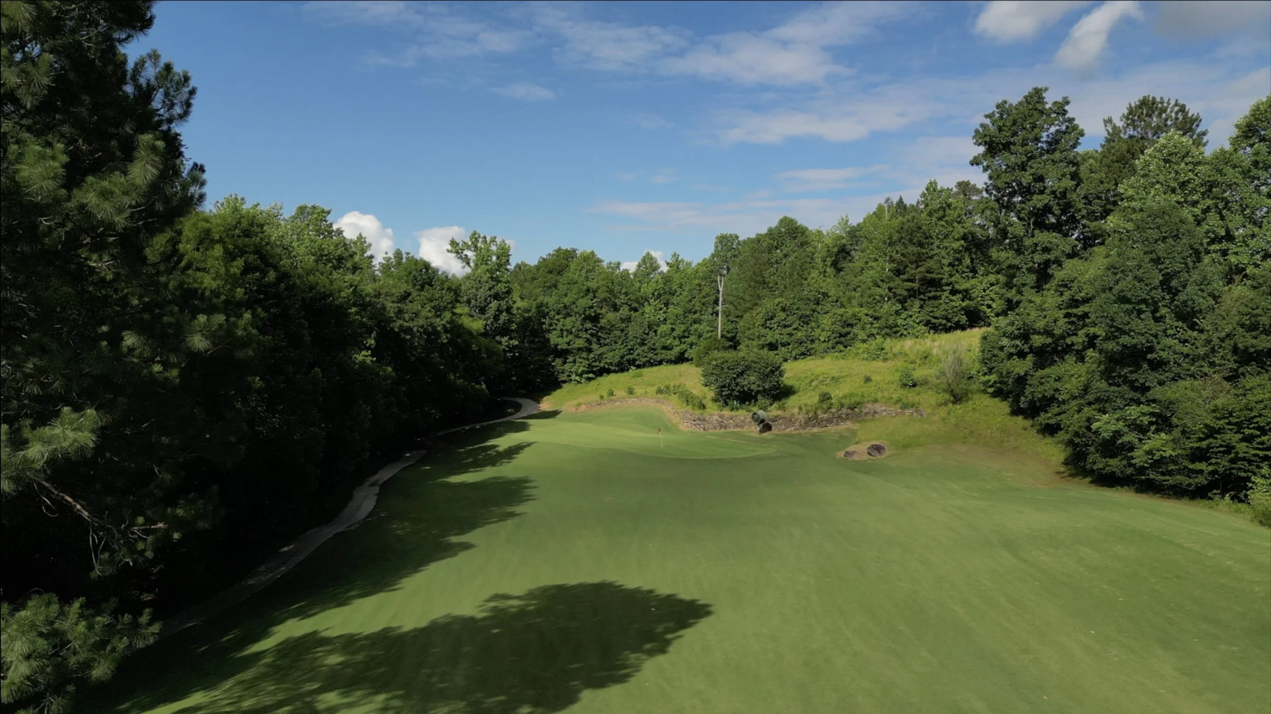 A scenic golf course fairway surrounded by tall green trees under a partly cloudy blue sky.