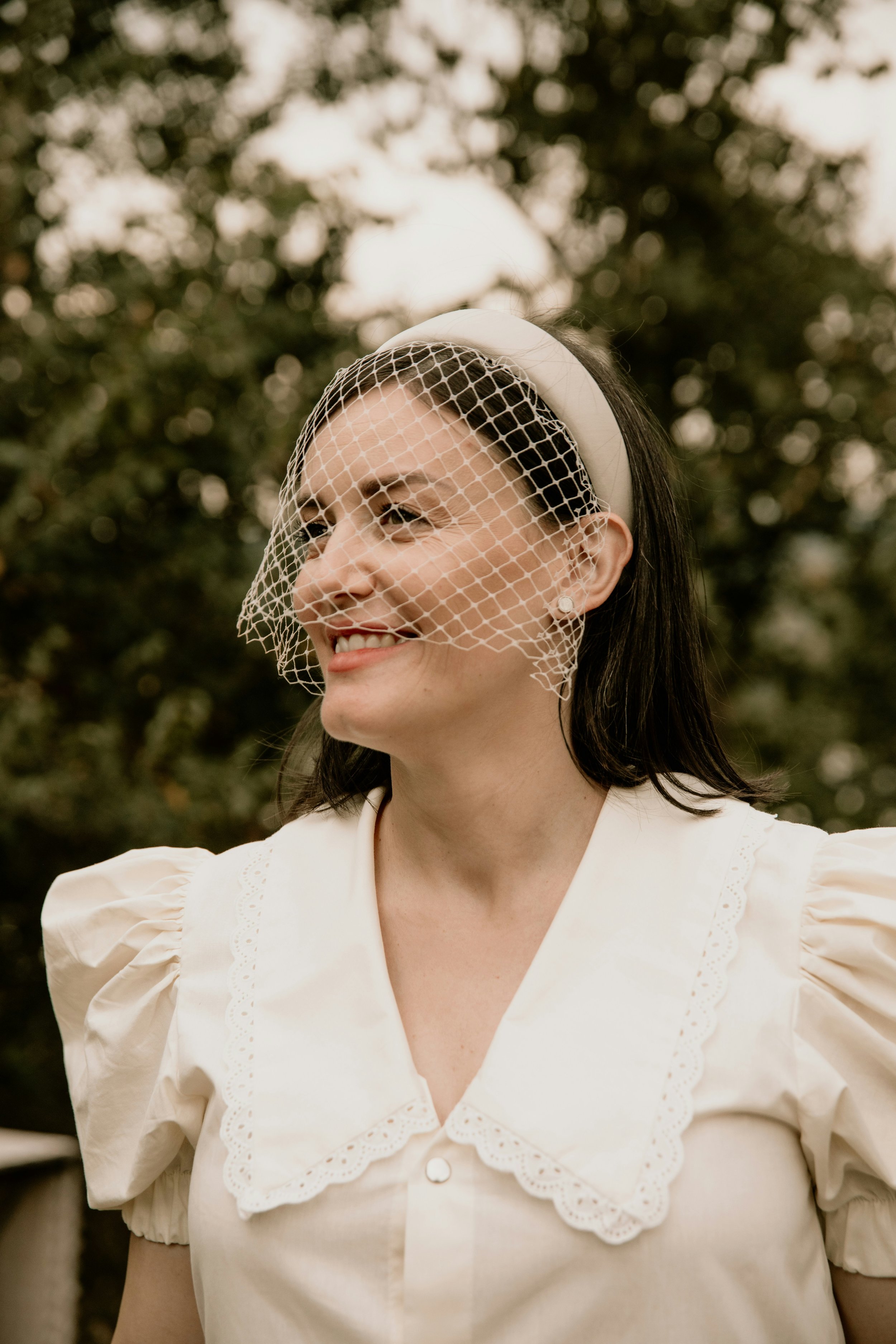 Woman smiling outdoors wearing a vintage white dress and a beige headband with a veil.