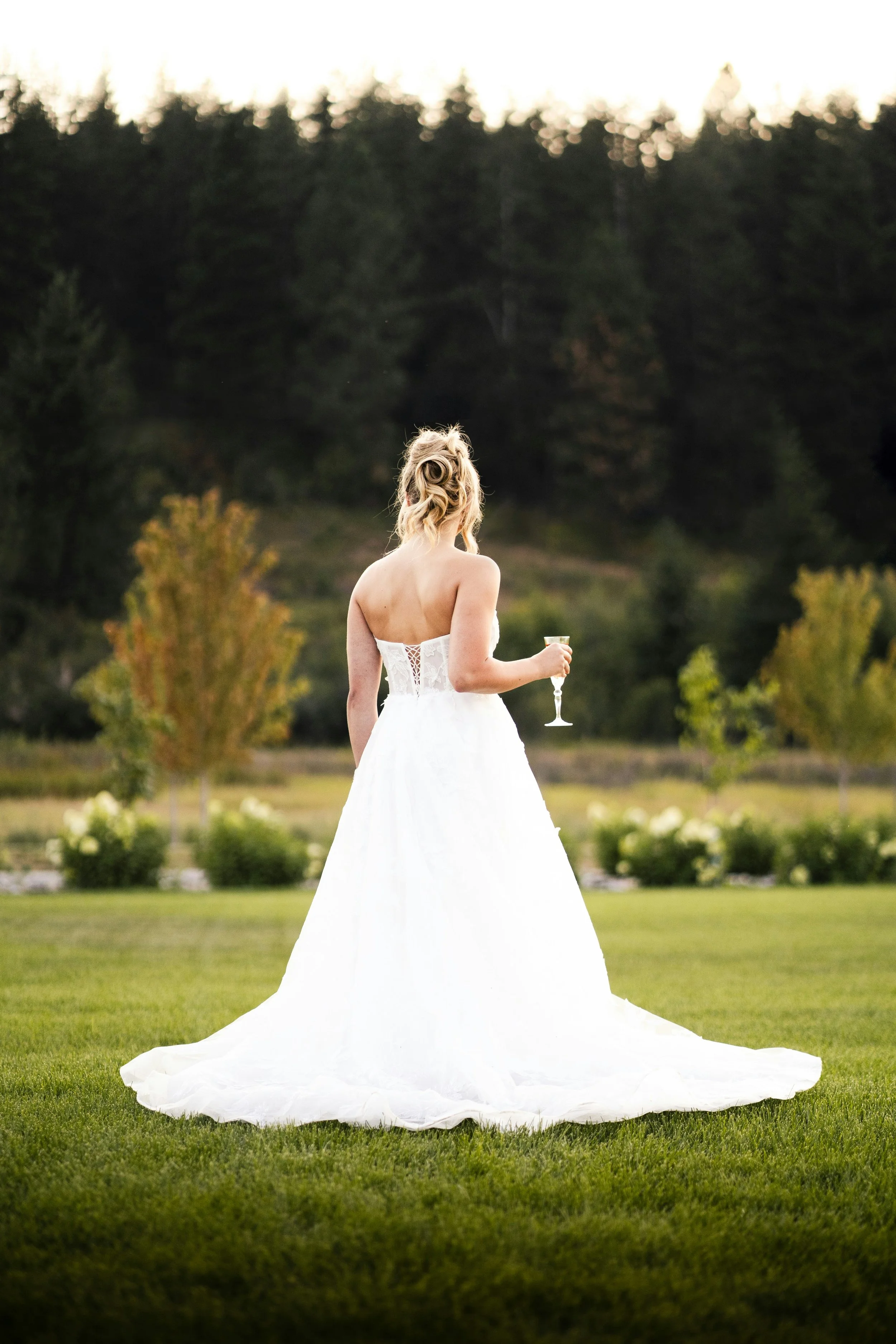 Bride in a white wedding dress holding a glass of champagne, standing on a lush green lawn with trees and a hillside in the background.