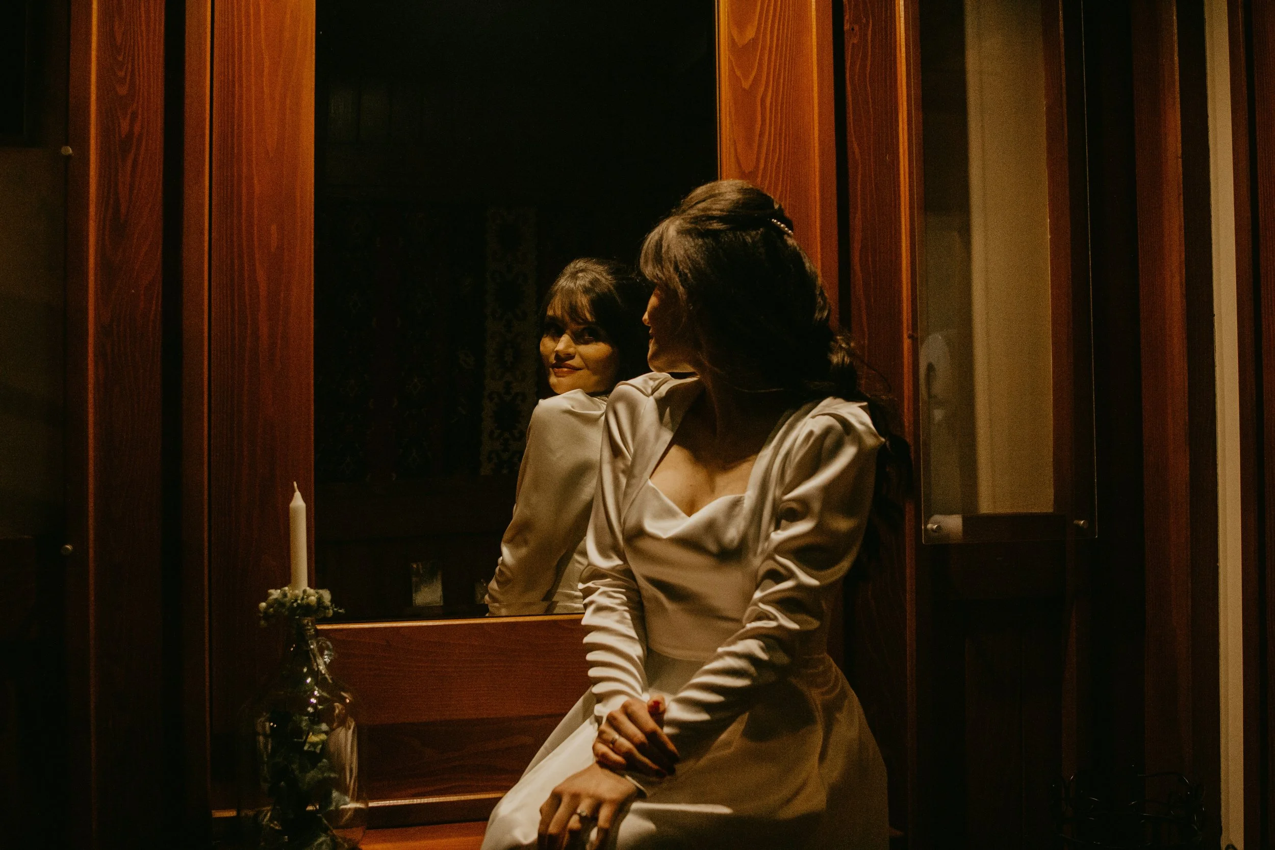 A woman in a cream-colored dress sitting on a bench, looking in a mirror that reflects her face. There is a candle on a stand next to her. The setting appears to be a dimly lit room with wooden paneling.