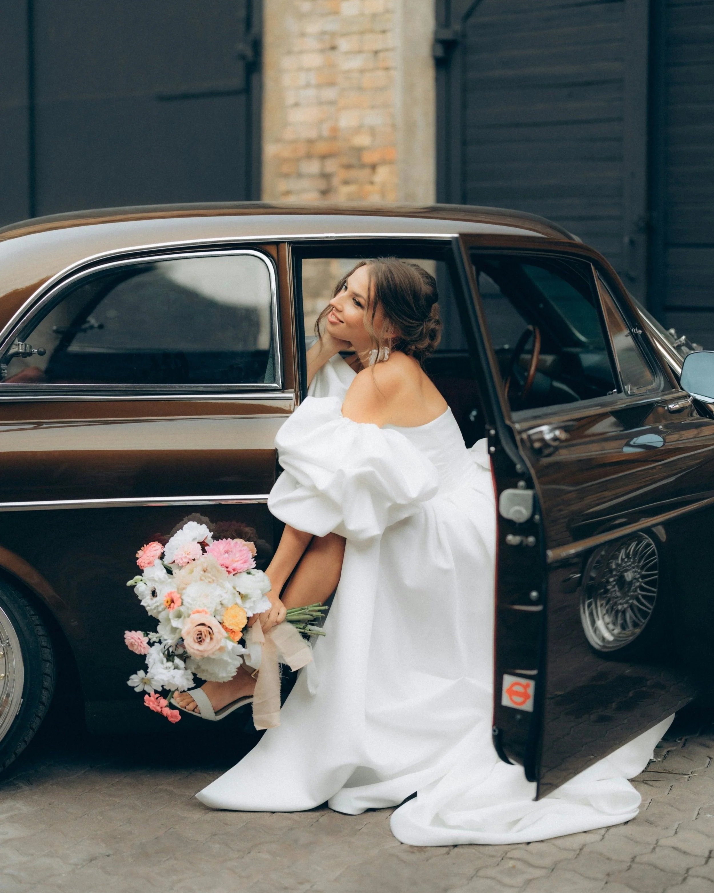 Bride in a white wedding dress holding a bouquet of pink and white flowers sits beside a vintage brown car with the door open.