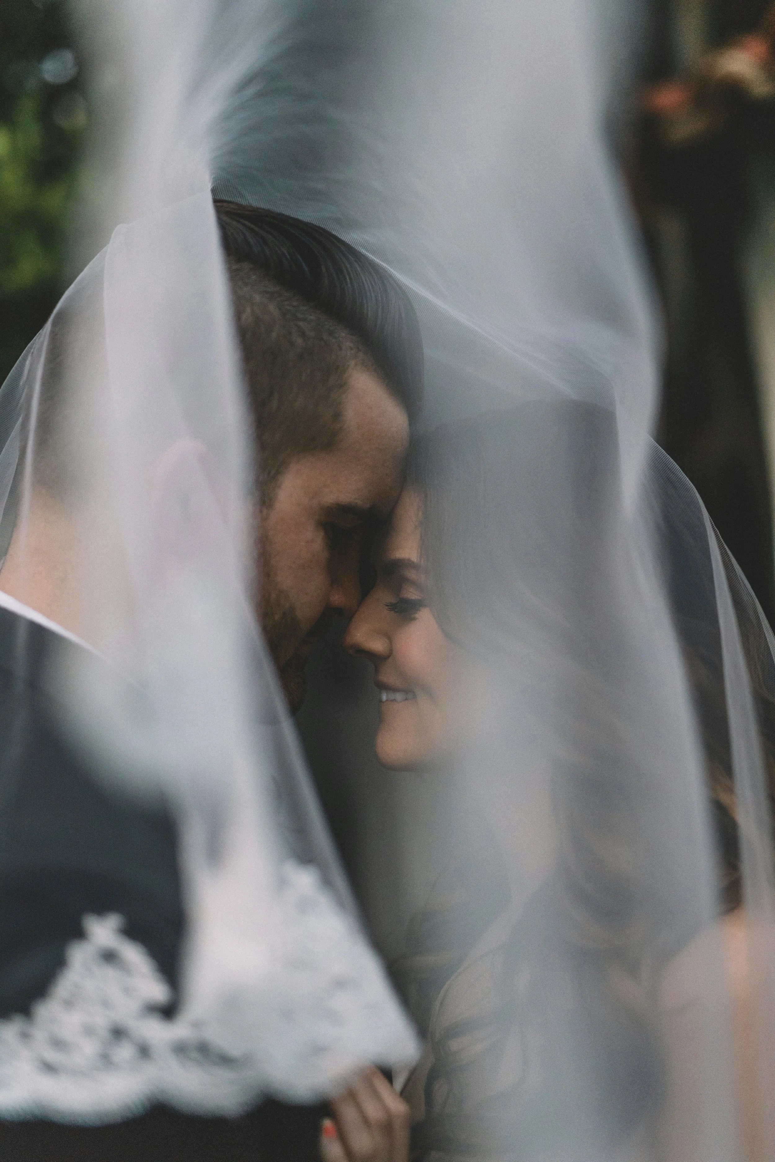 A couple close together under a veil, smiling and touching noses in an intimate moment.