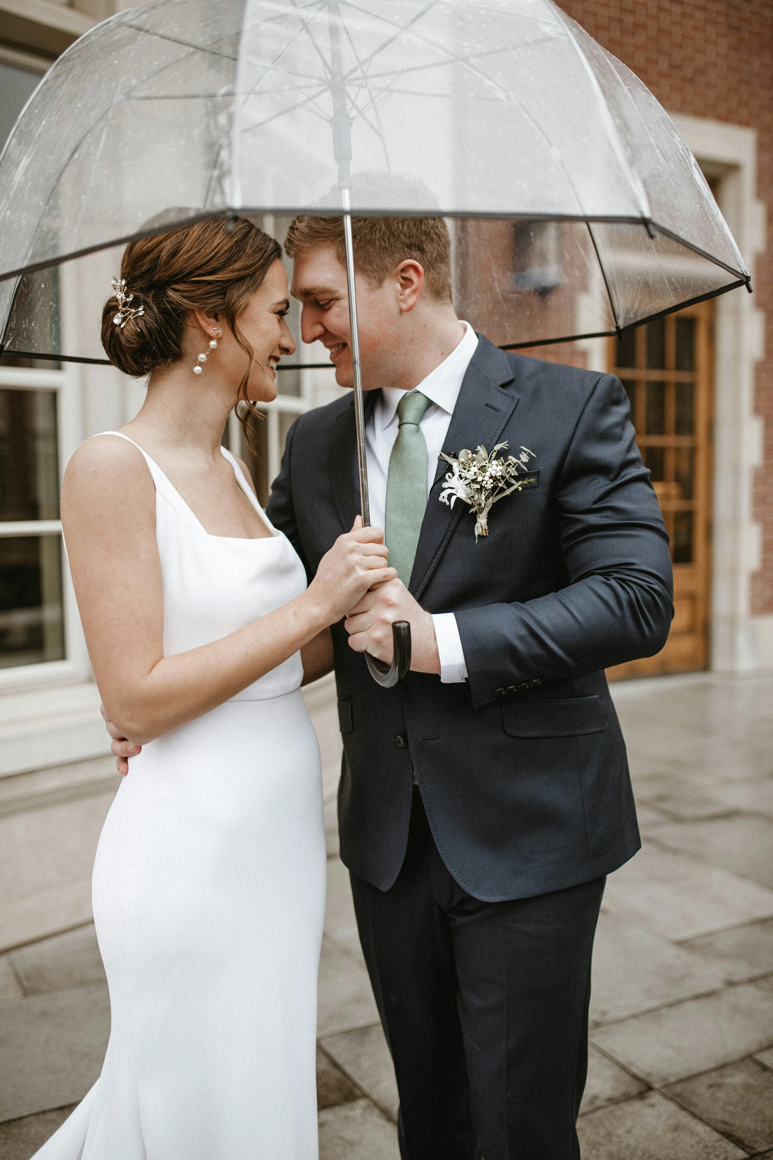 Bride and groom under a transparent umbrella, smiling and leaning close together outside a building.