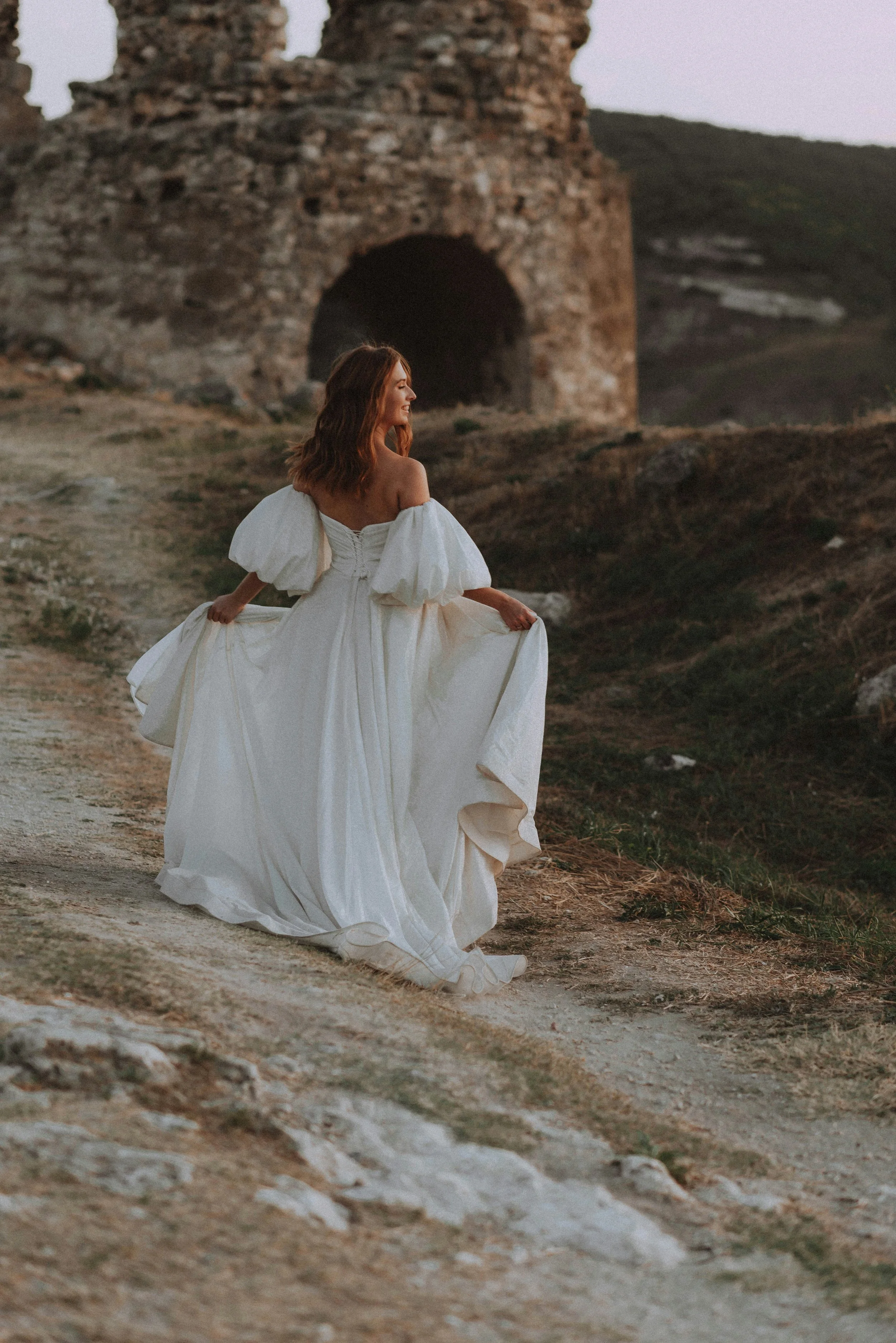 A woman in a white wedding dress walking outdoors near ancient ruins during sunset.