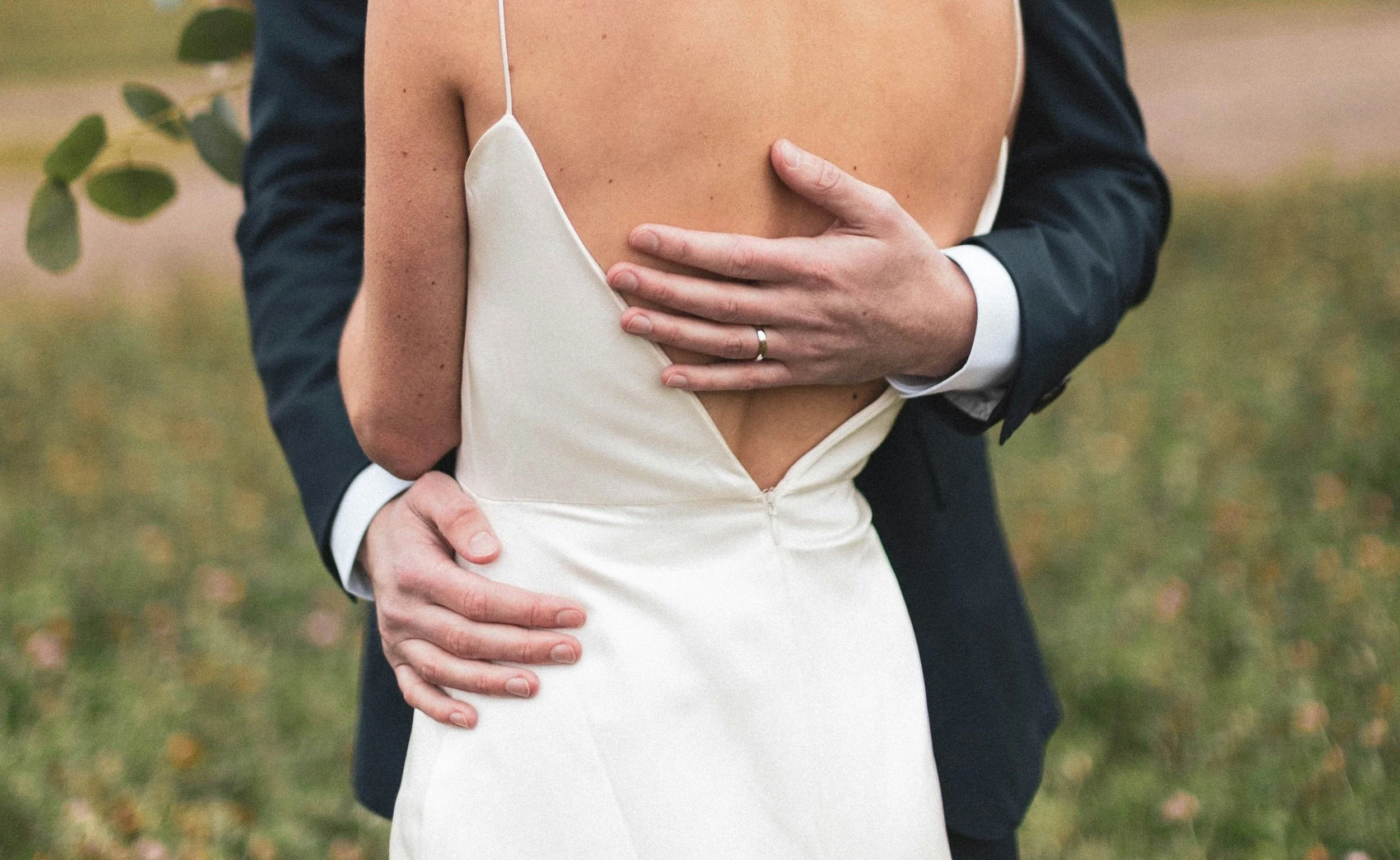Close-up of a bride in a wedding dress and groom in a suit embracing outdoors, focusing on their torsos and hands.