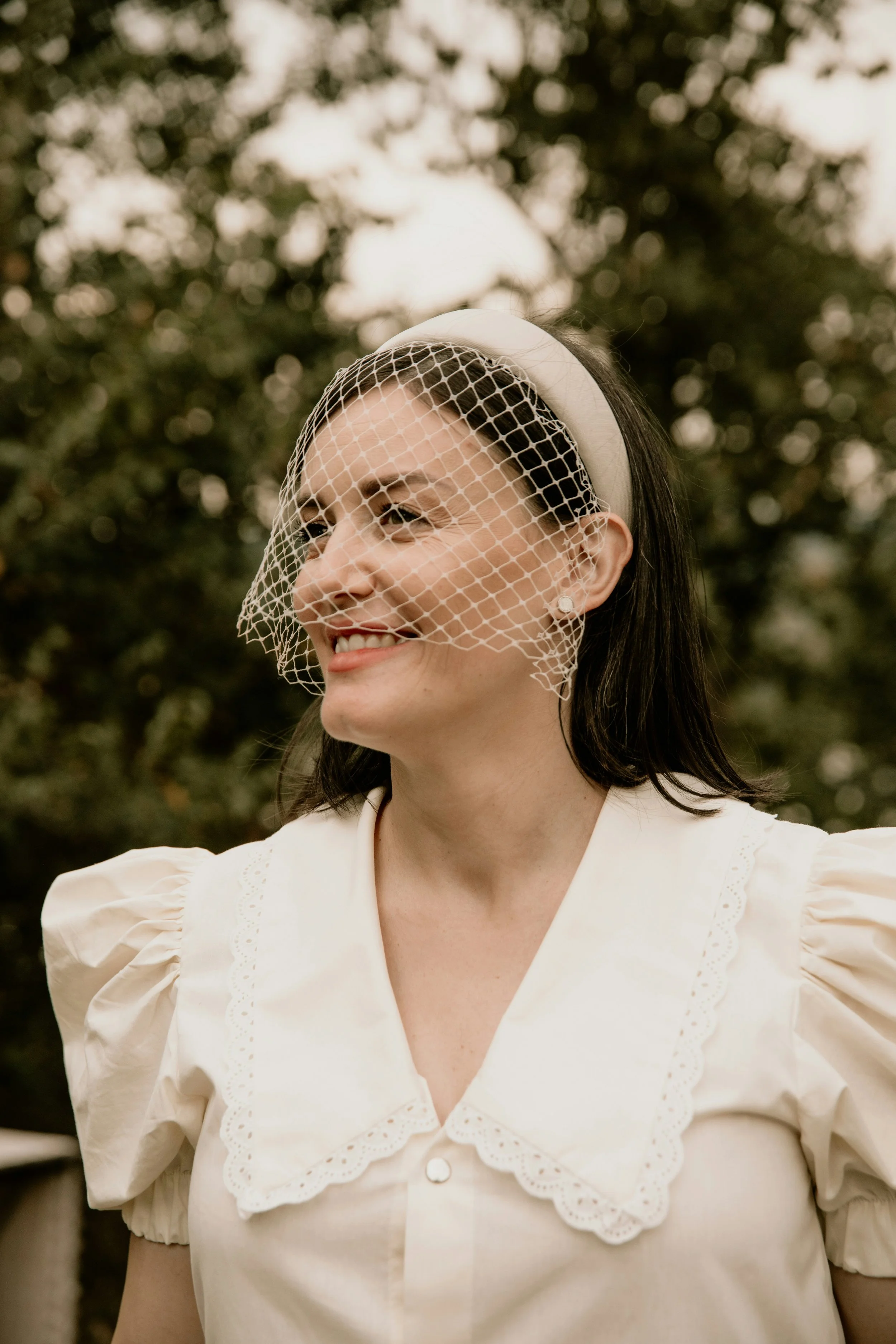 A woman with dark hair, smiling, wearing a vintage white dress with a scalloped collar and puffed sleeves, a pearl earring, and a veil headband, outdoors with trees in the background.