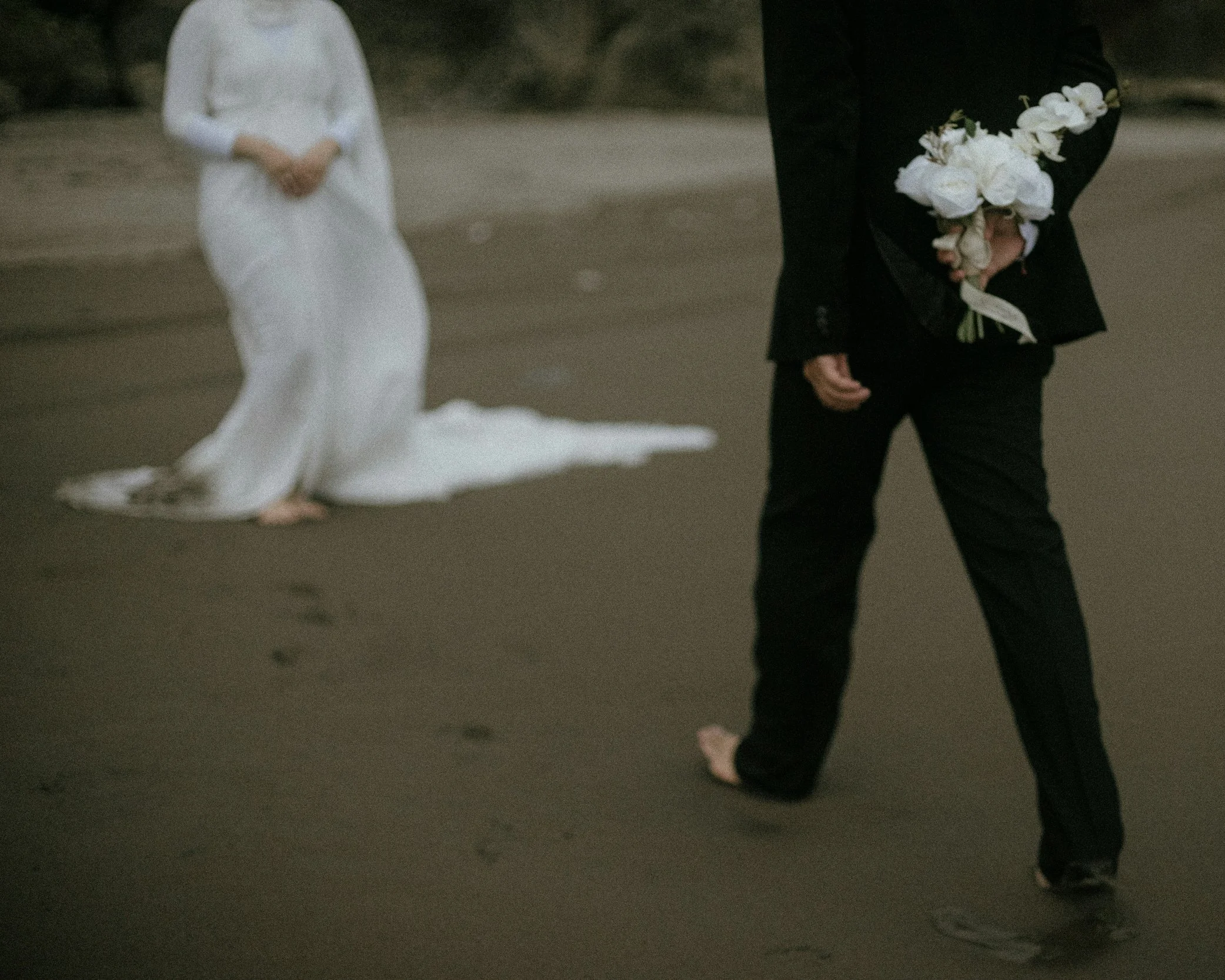 A person in a black suit holding a bouquet of white flowers standing on a beach, with a woman in a white wedding dress kneeling in the background.