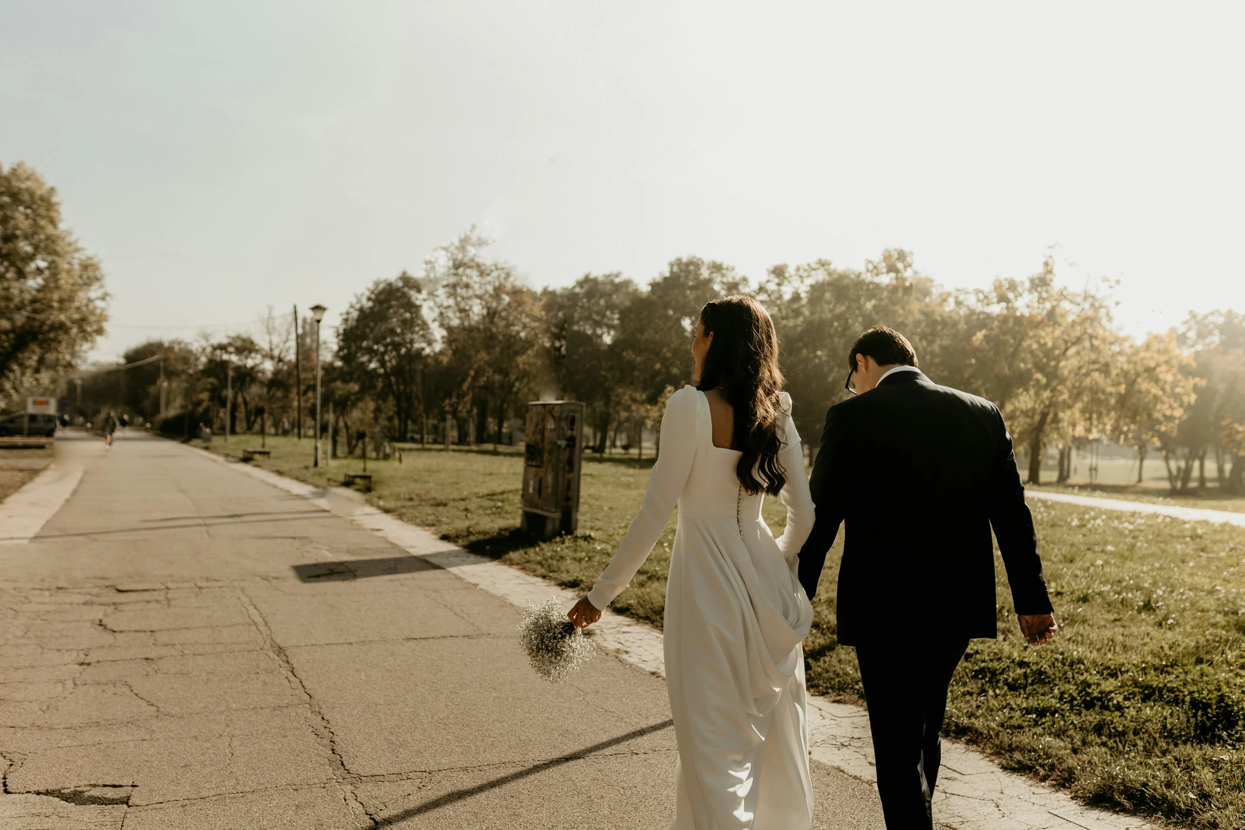 A bride and groom walking hand in hand in a park during sunset
